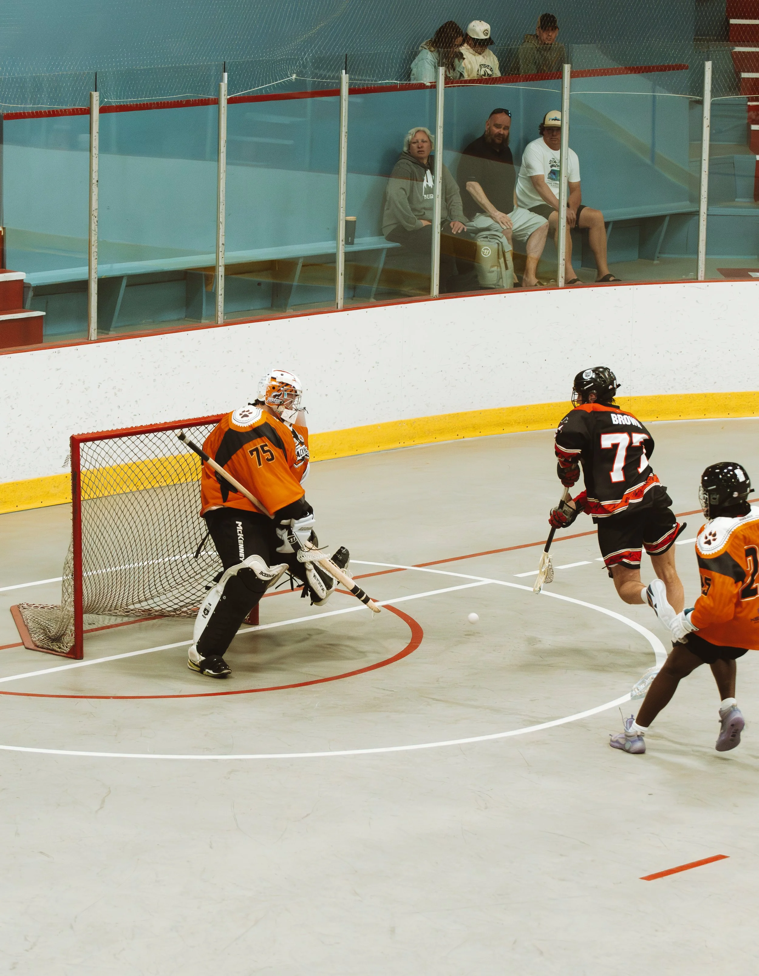 Ice hockey game in progress with players on the ice, including a goalkeeper in black and white gear, and players in black and orange jerseys, near the goal. Spectators sit behind protective glass, watching the action.