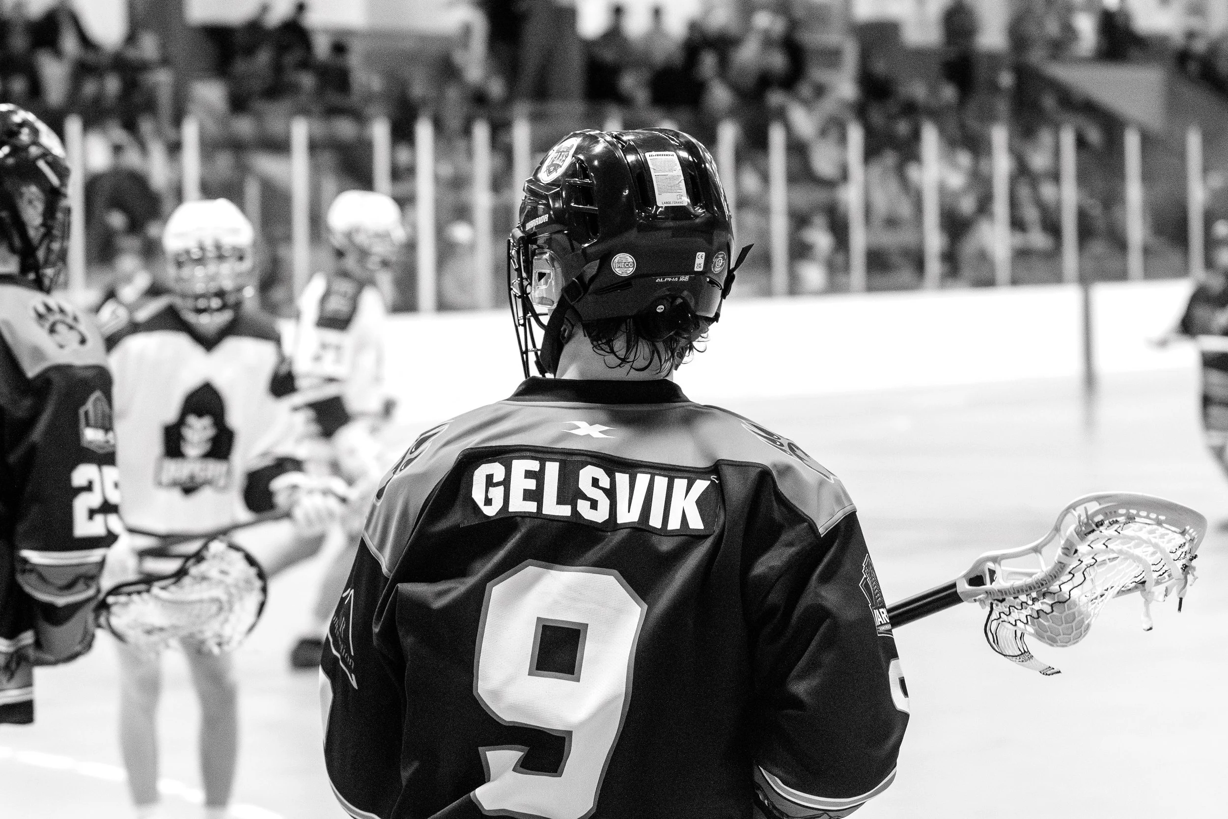 Black and white photo of box lacrosse player Gelsvik, number 9, photographed from behind holding his stick on the arena floor with other players visible near the boards.