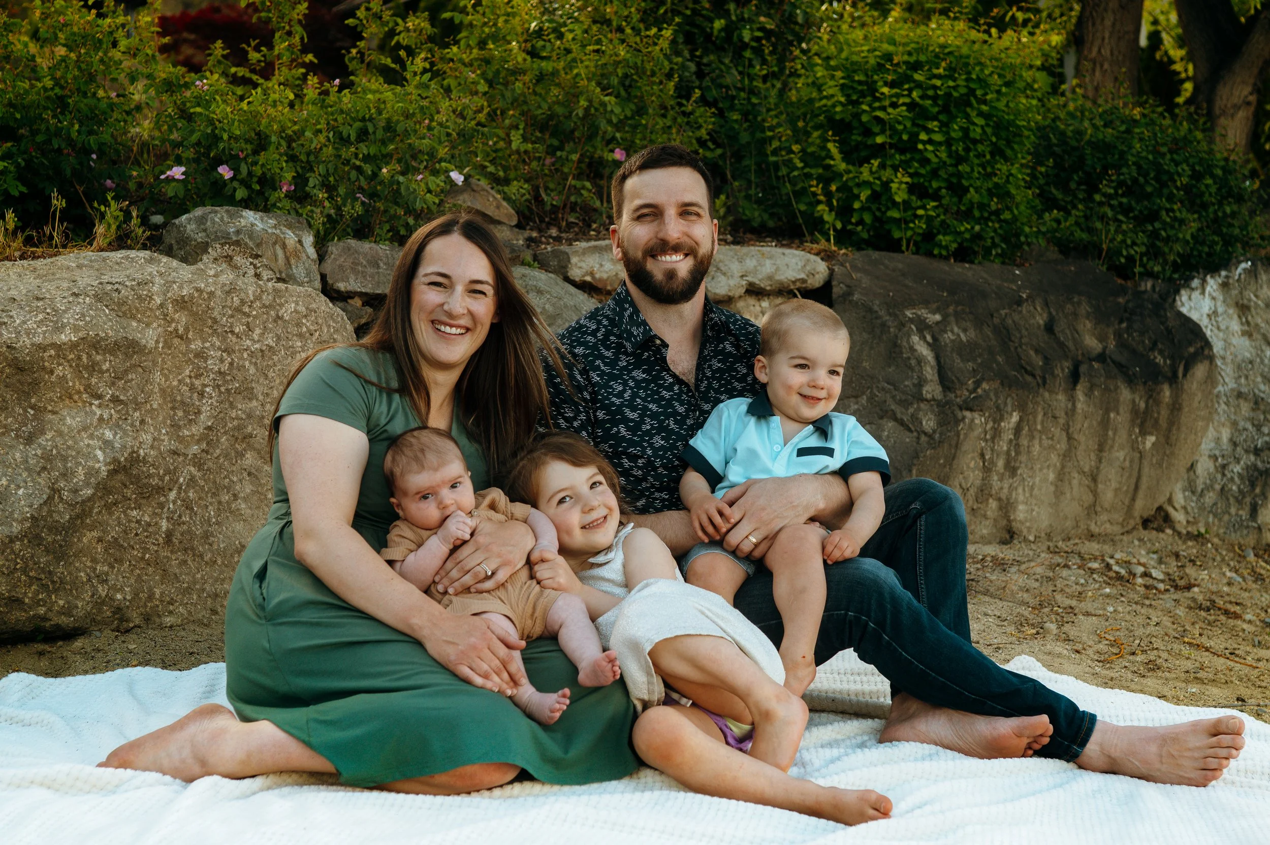 Family of six sitting on blanket outdoors with rocks and green bushes behind them, smiling.