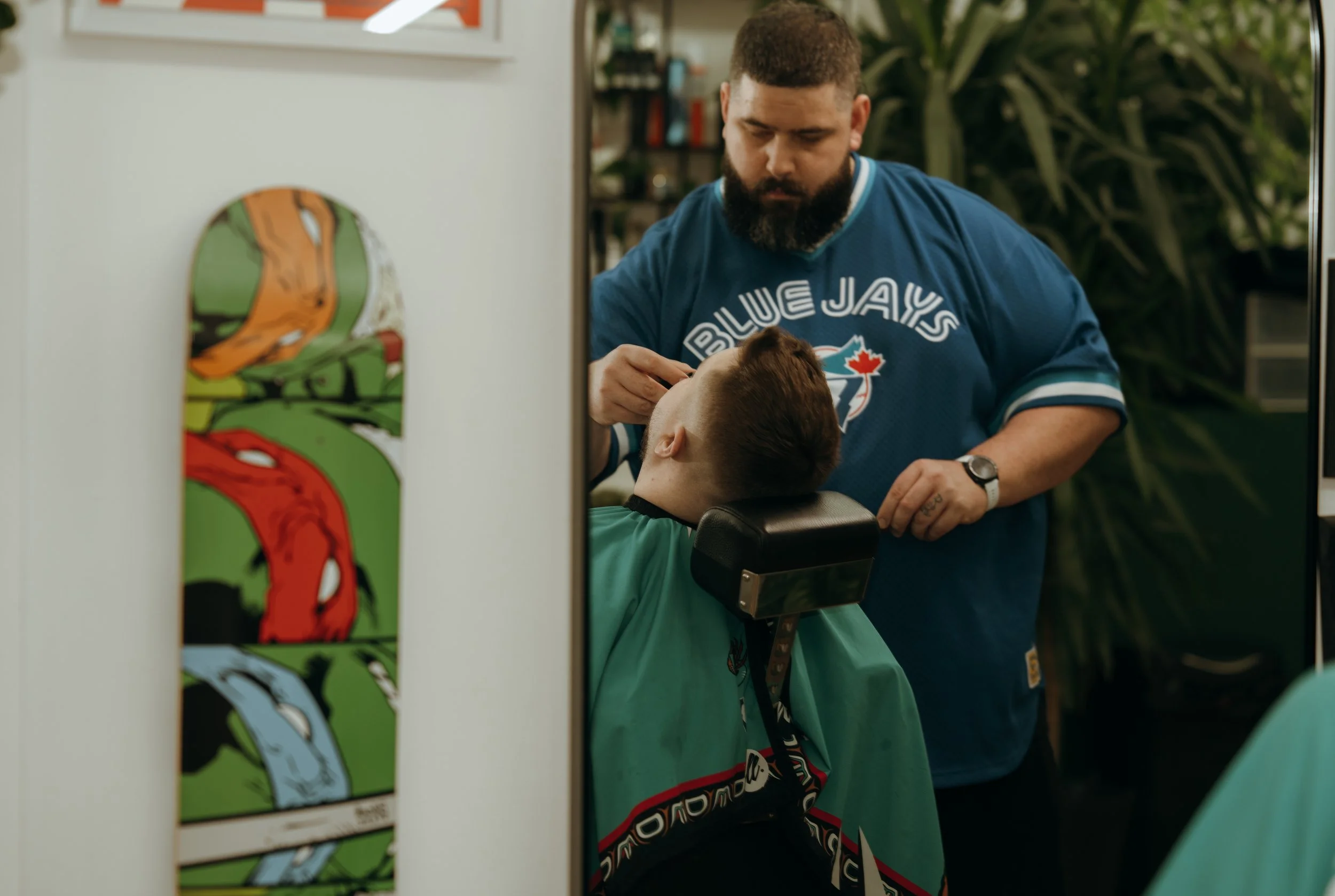 A man getting a haircut at a barber shop, wearing a Toronto Blue Jays T-shirt, while the barber trims his hair. A skateboard with a colorful design hangs on the wall nearby.