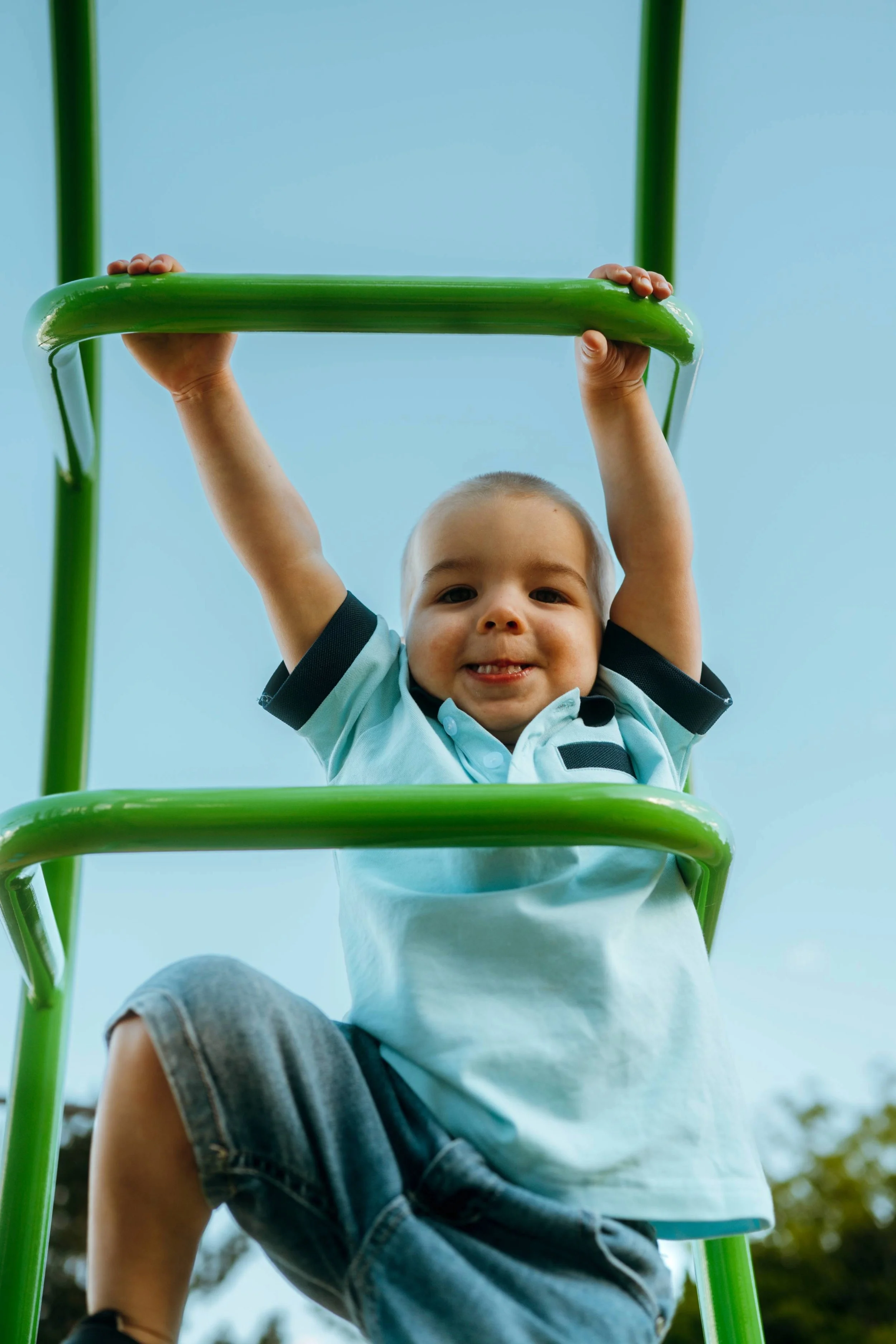 Child climbing on a green jungle gym, smiling and looking at the camera, outdoors with trees and sky in the background.