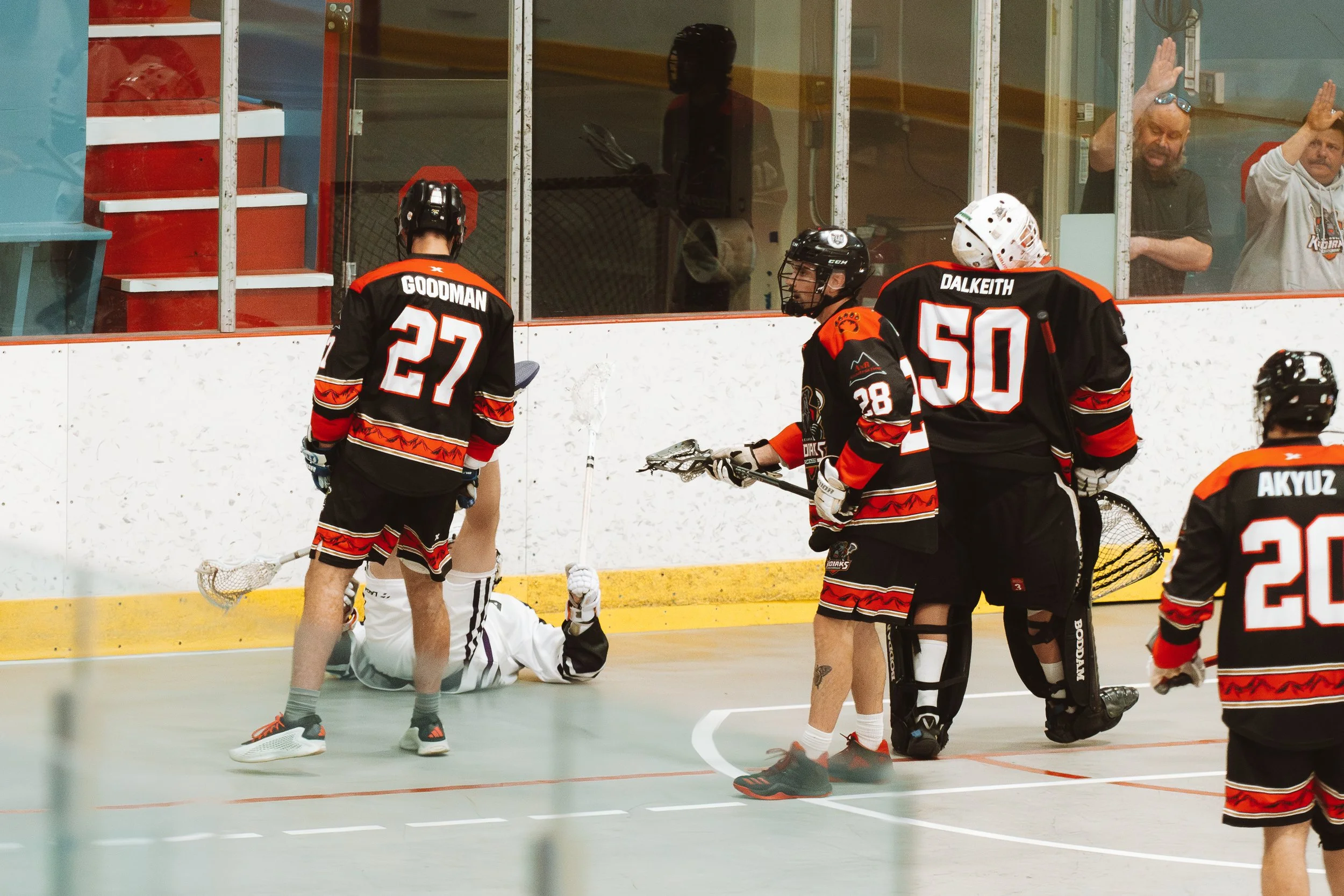Kodiaks box lacrosse players Goodman, Dalkeith, and teammates gather near the boards during a game while an opposing player in white lies on the floor from a hit by Goodman. 