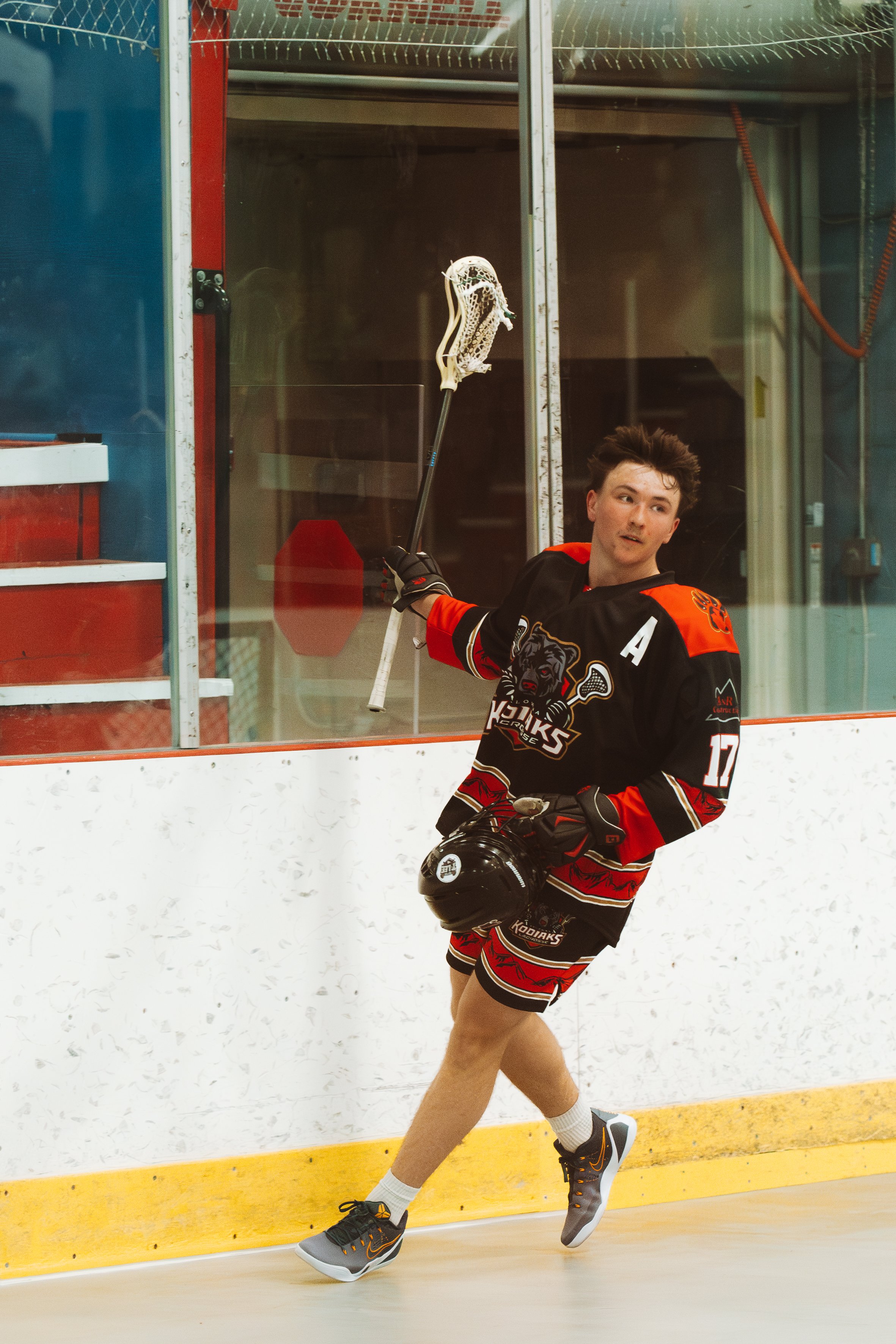 Box lacrosse player wearing a black Kodiaks jersey with number 17 and an assistant captain patch, walking along the arena boards while carrying a helmet and raising his lacrosse stick.