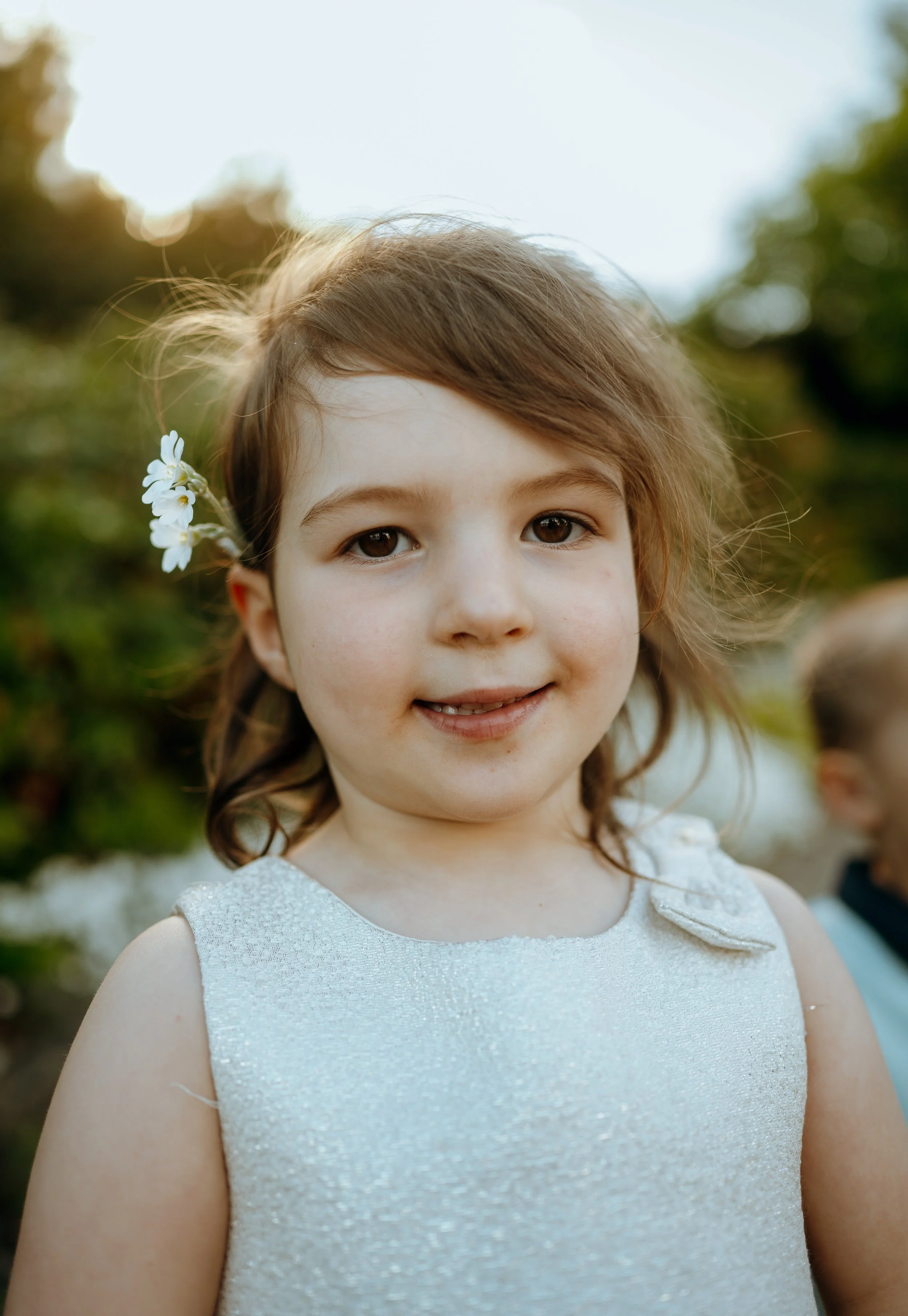 A young girl with brown hair, wearing a white dress, with flowers tucked behind her ear, smiling outdoors with blurred greenery in the background.