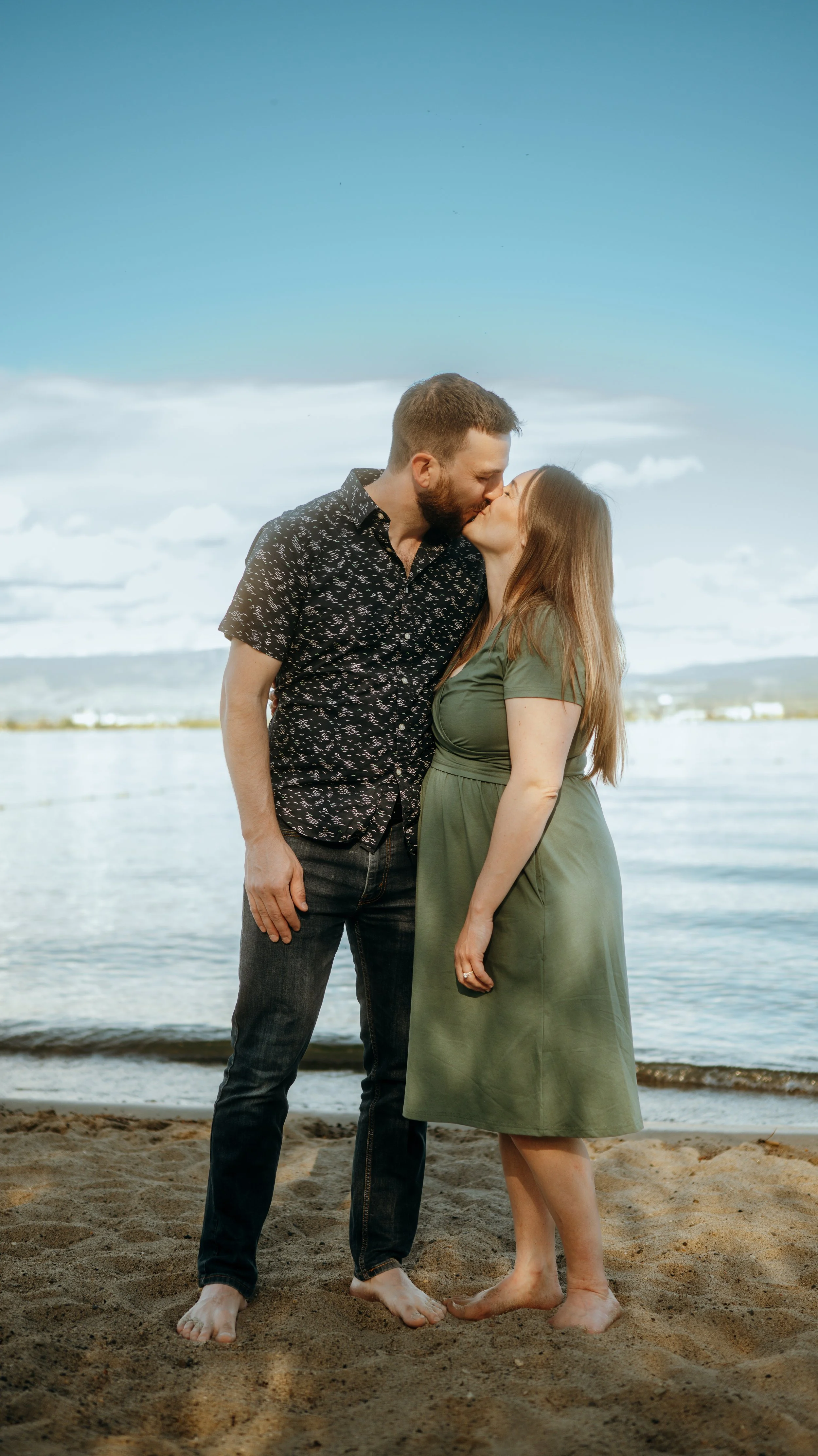 A couple sharing a kiss at the beach, standing barefoot on sand with their foreheads touching, water and sky in the background.