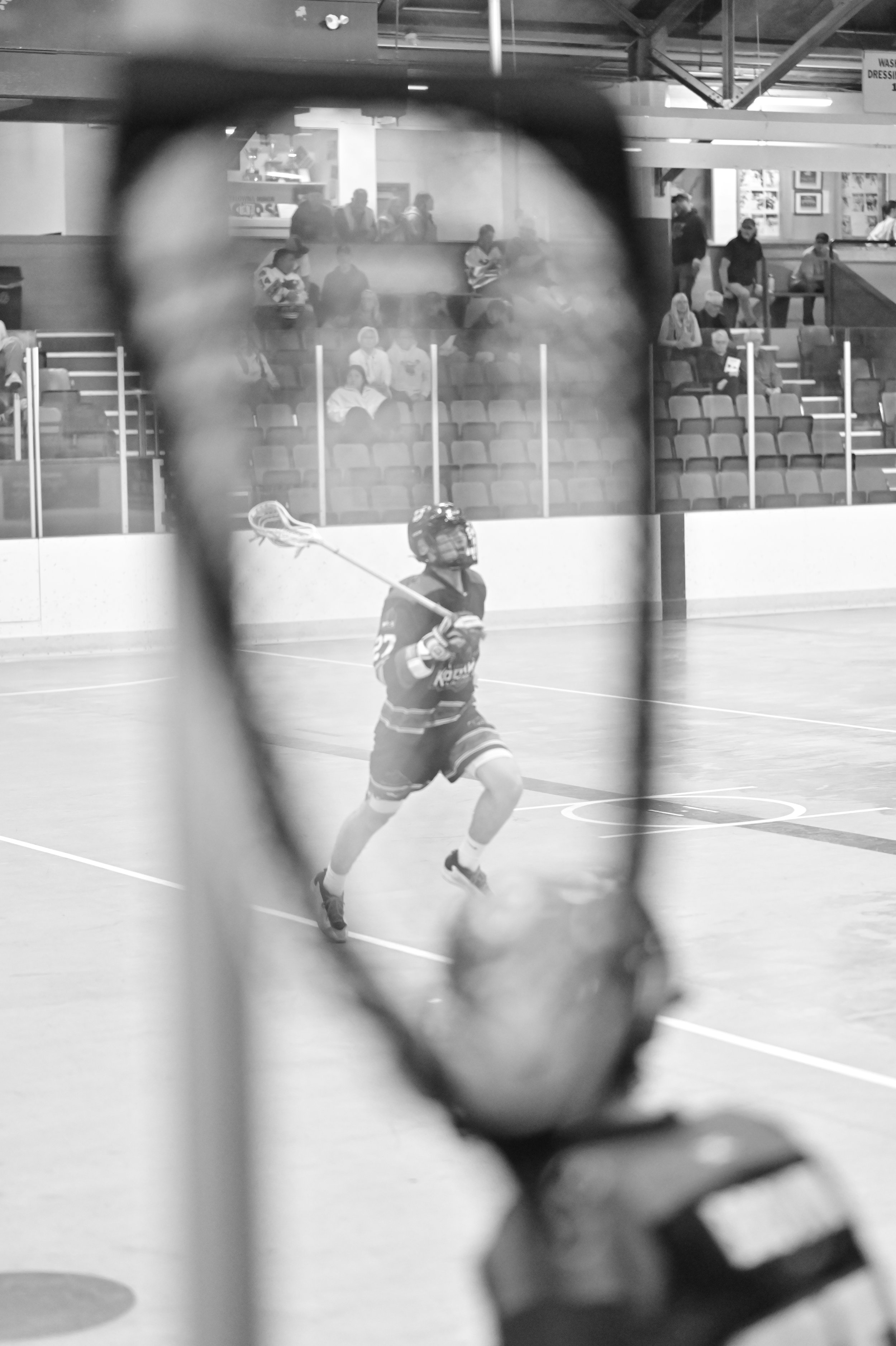 Black and white photograph of an indoor hockey game, viewed through a fence. A player in hockey gear is in the foreground, skating on the ice. Spectators are seated in the background, watching the game.