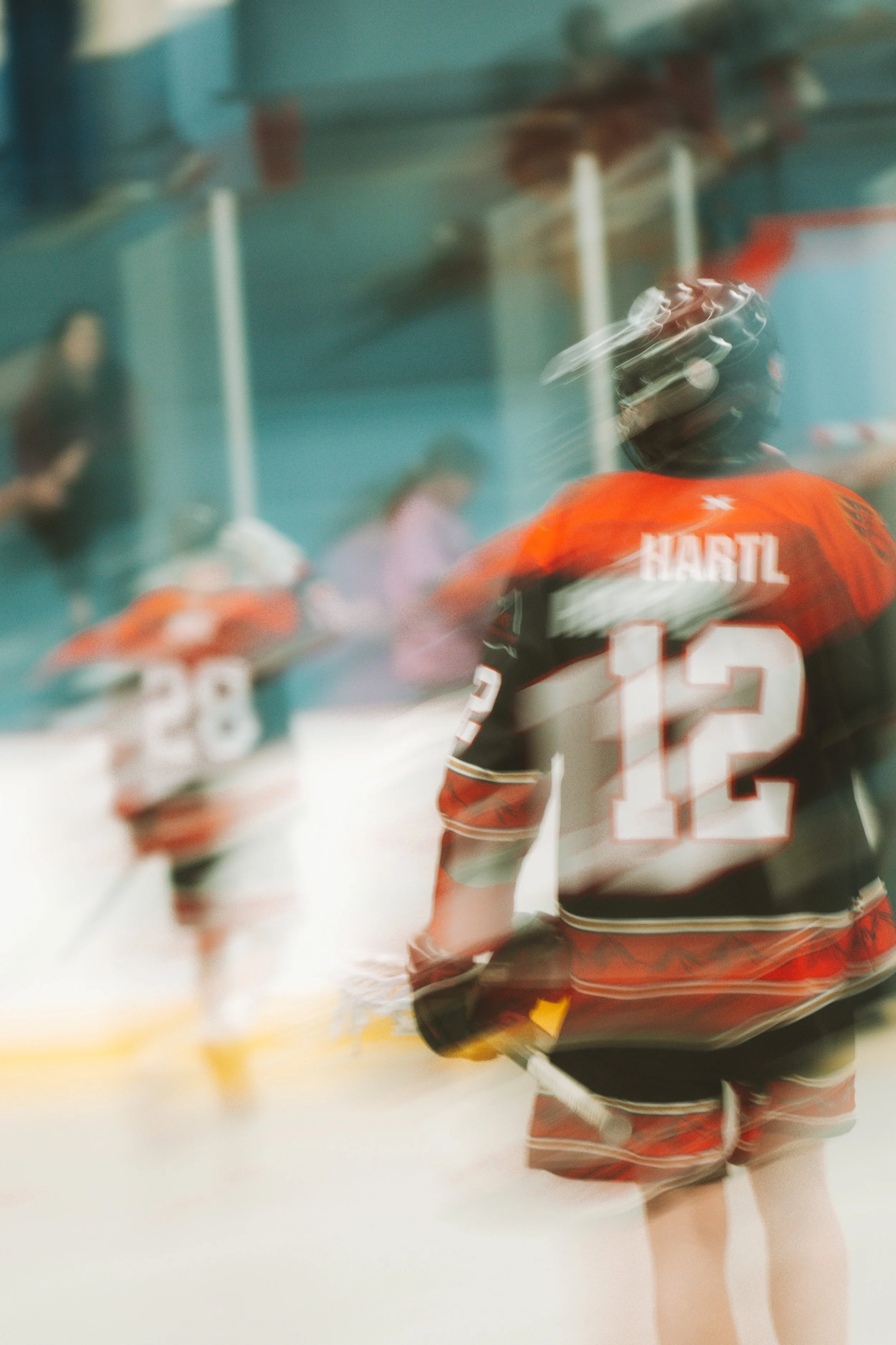 Motion-blurred box lacrosse player wearing an orange and black jersey with the name Hartl and number 12, running on the arena floor with a teammate visible in the background.