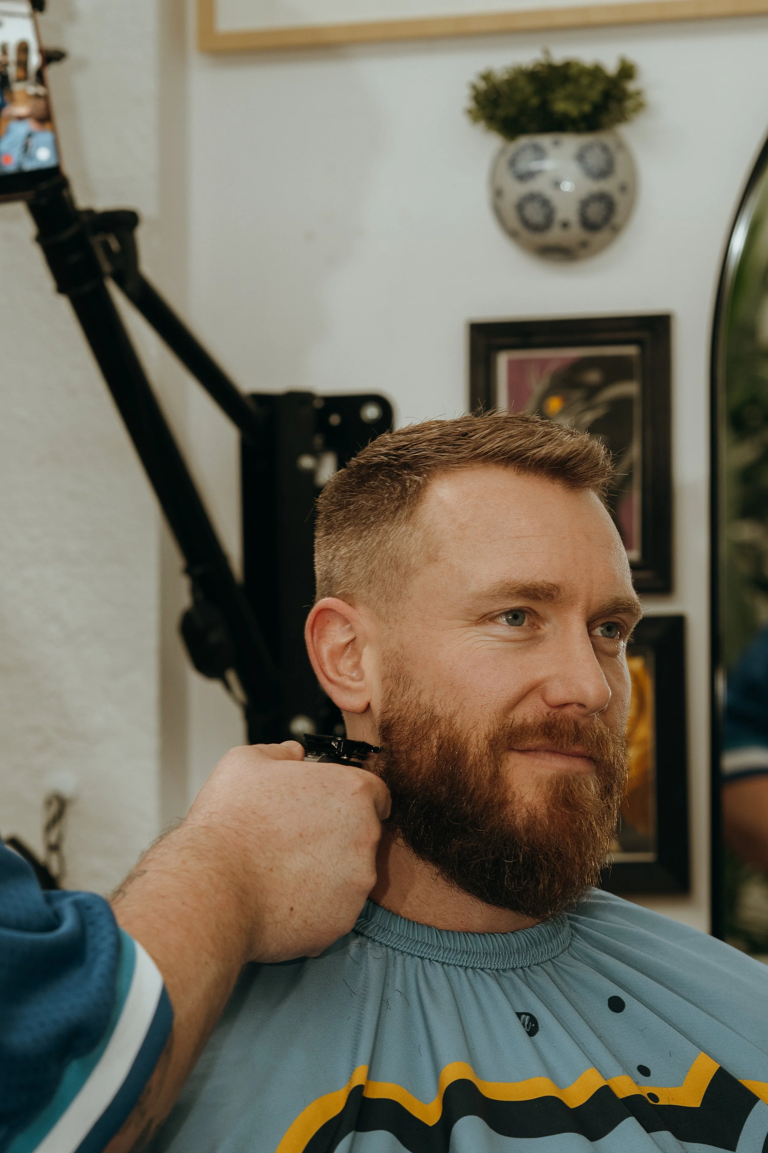 A man with a beard gets a haircut at a barber shop, sitting in a barber's chair while a barber shaves his neck with clippers.
