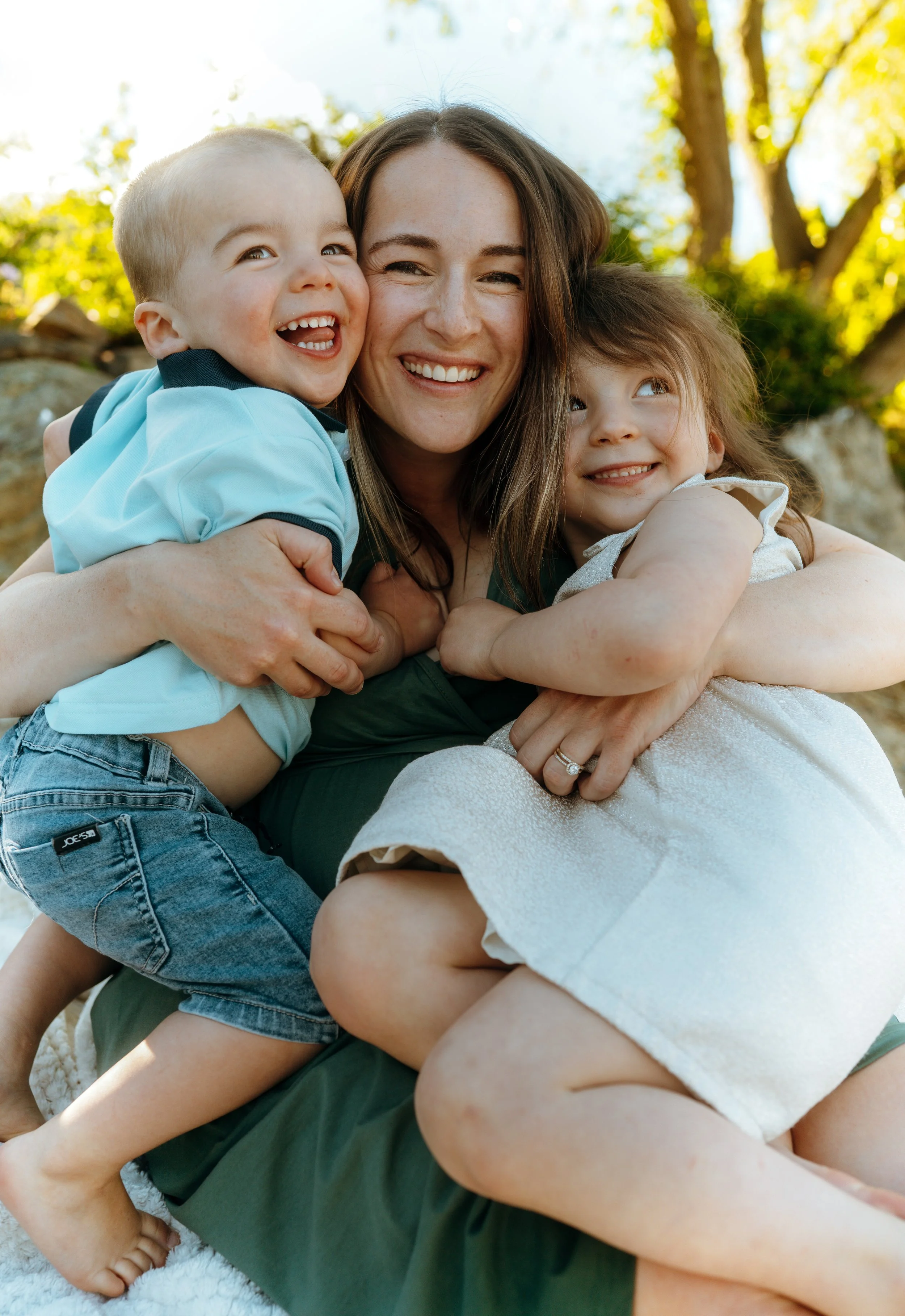 A woman smiling and hugging two children outdoors during daytime, surrounded by trees with green leaves.