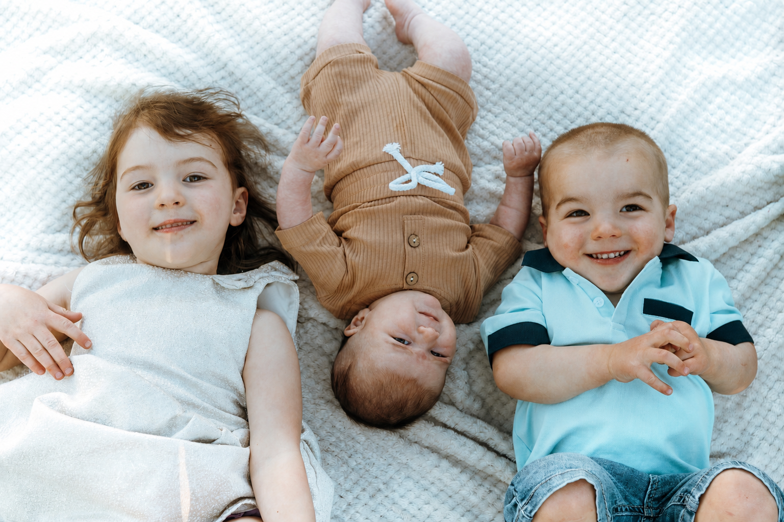 Three young children lying on a soft white blanket, smiling and looking at the camera, with the baby in the middle upside down.