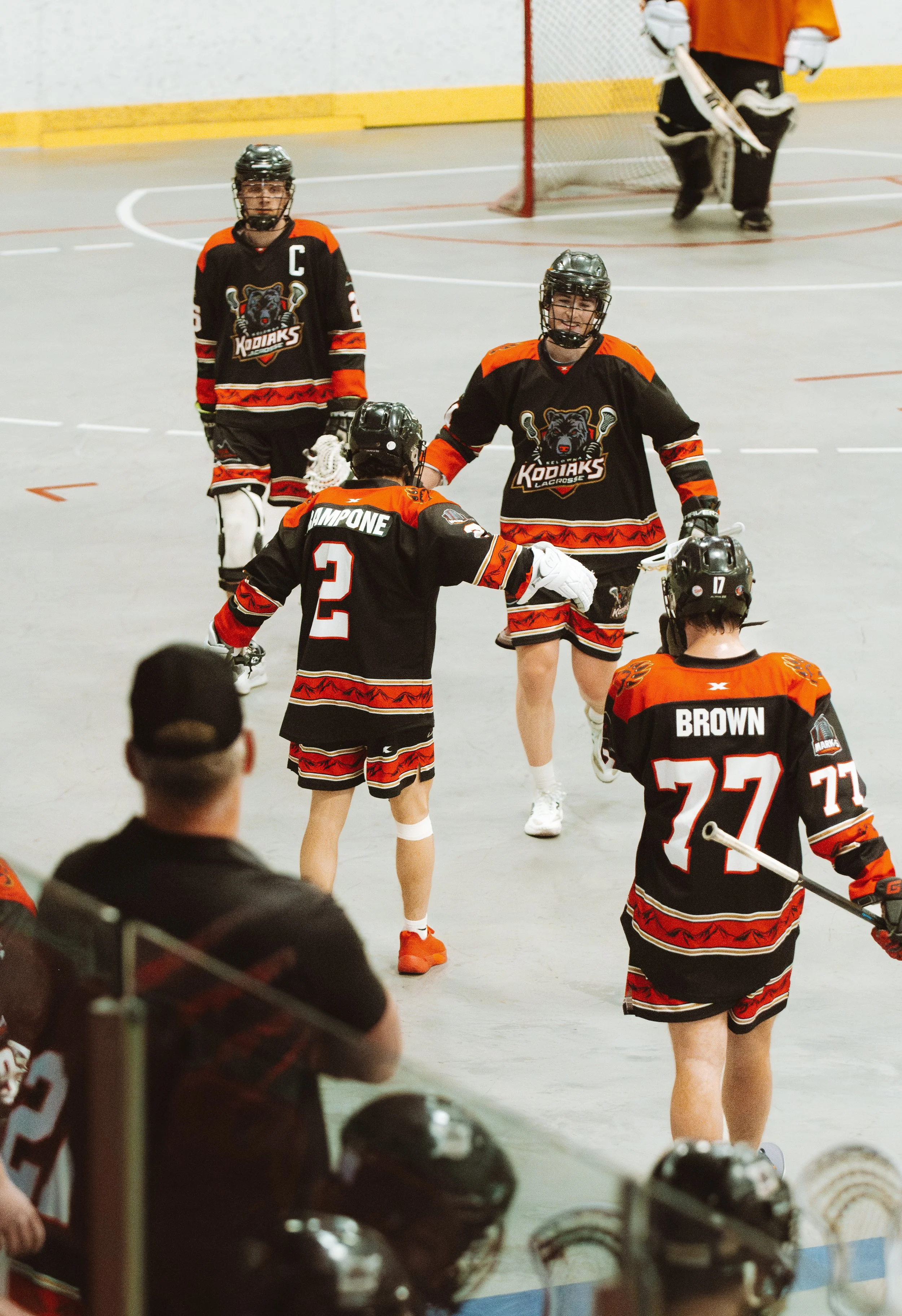 Hockey players celebrating on the rink, wearing black jerseys with red, orange, and white accents, and a logo of a bear with hockey sticks. One player is wearing a captain's 'C', and some players are shaking hands or smiling. Helmets and hockey equip
