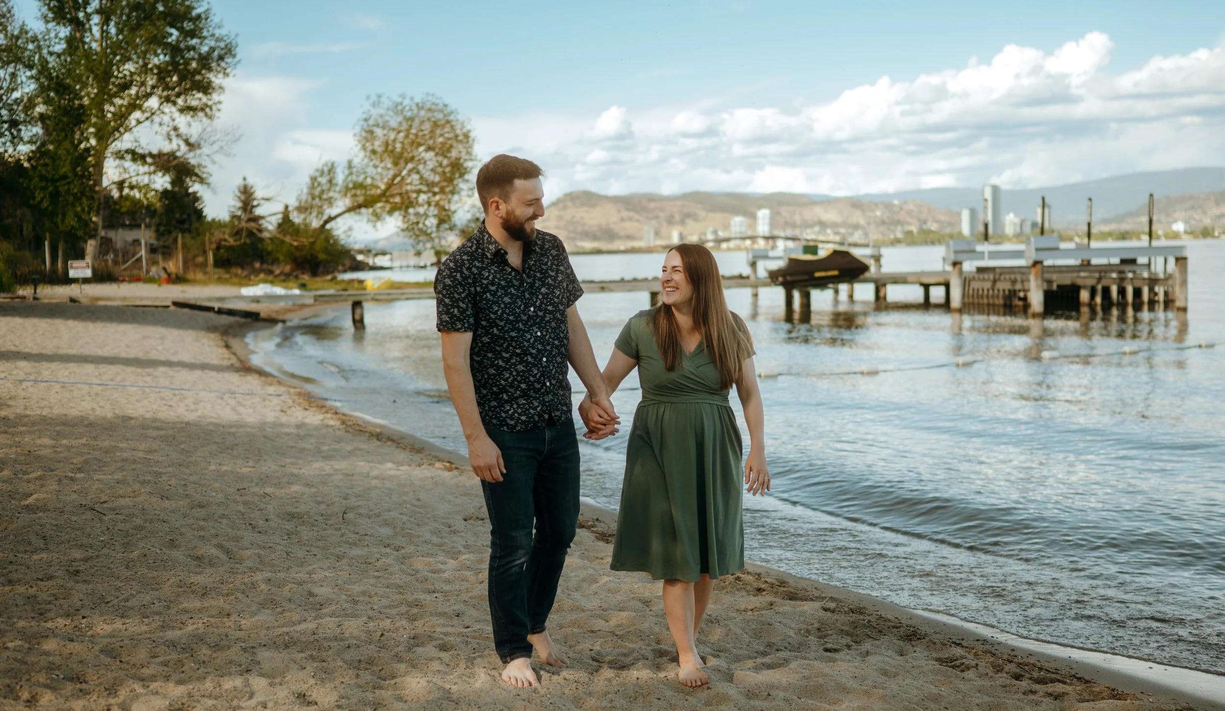 A happy couple walking barefoot along a sandy beach near a body of water, holding hands and smiling at each other, with trees, buildings, and mountains in the background.