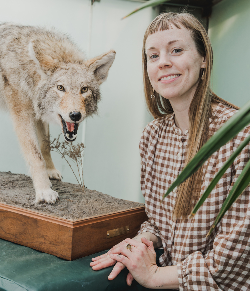 Artist Maggie Dimmick smiles in front of a taxidermy coyote at a museum