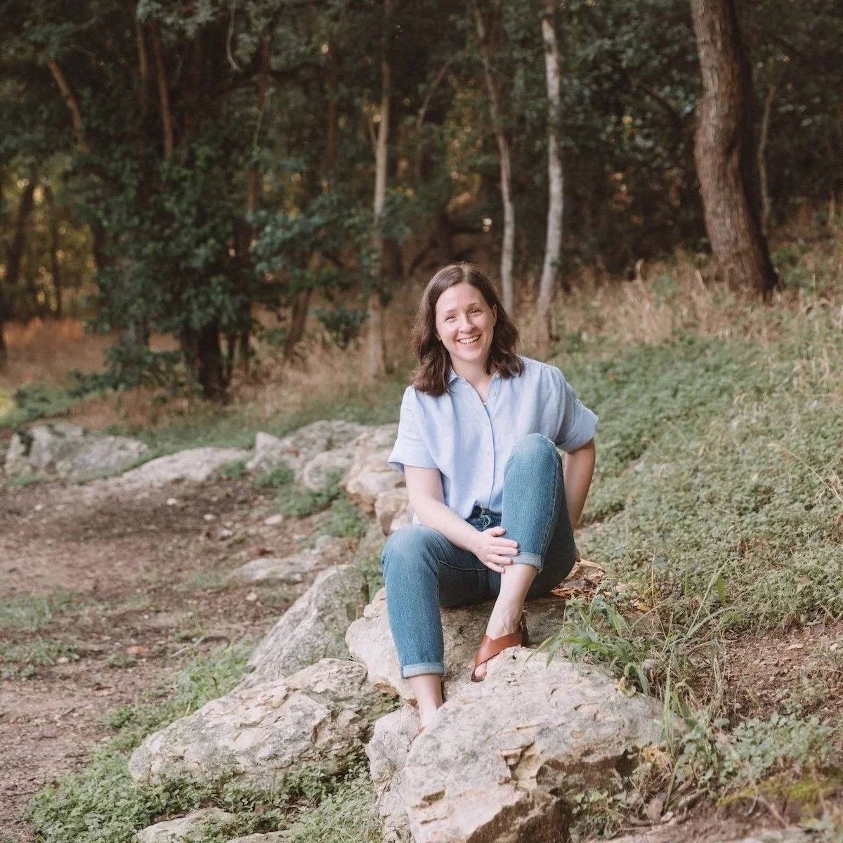 Sister Seasons' founder Rebecca Magee sits smiling on a rock with a forest behind her.