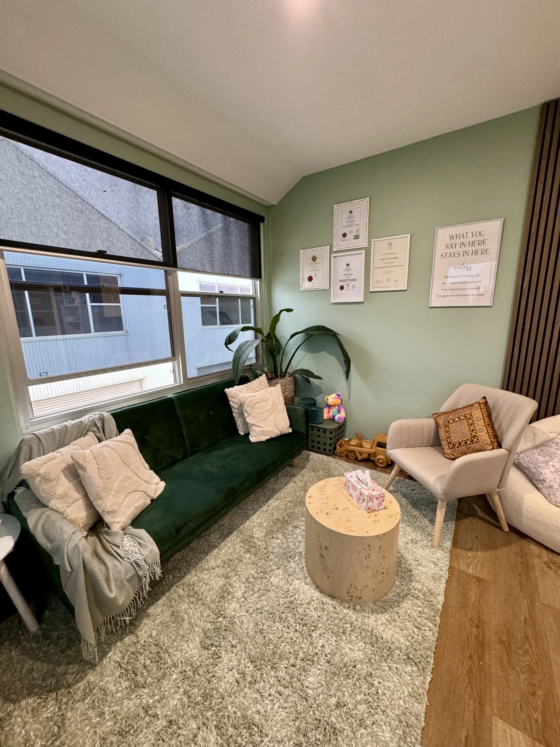 Cozy living room with a green velvet couch, beige pillows, throw blanket, circular beige coffee table, cream armchair with decorative pillow, large window, framed certificates on a mint wall, striped dark wood accent wall, plush rug, potted plant, toys, and tissue box.