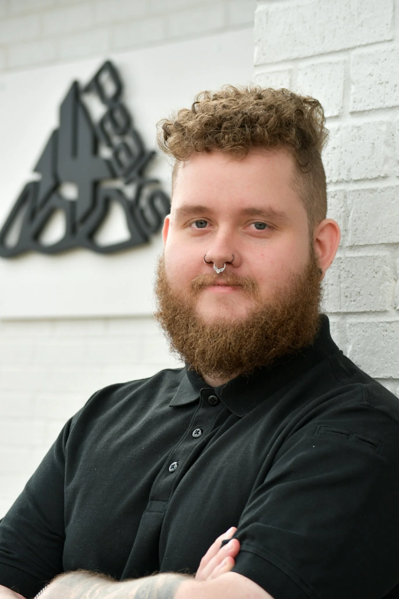 A young man with curly hair and a beard, wearing a black shirt, leans against a white brick wall with a black abstract art piece in the background.