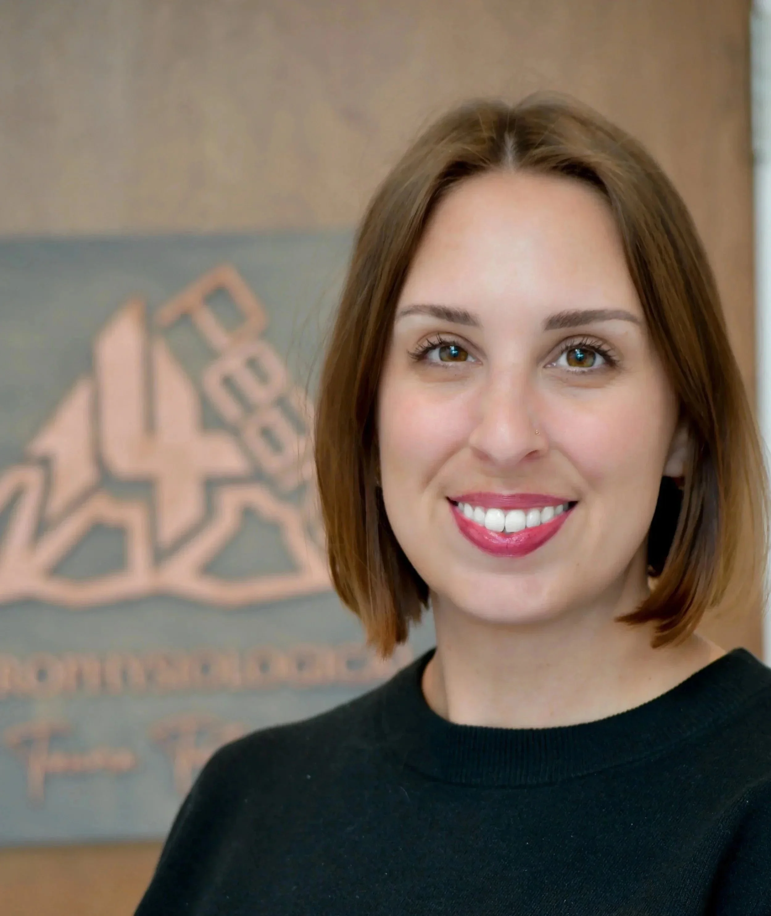 A smiling woman with shoulder-length brown hair, wearing a black top, standing indoors with a sign or artwork in the background.
