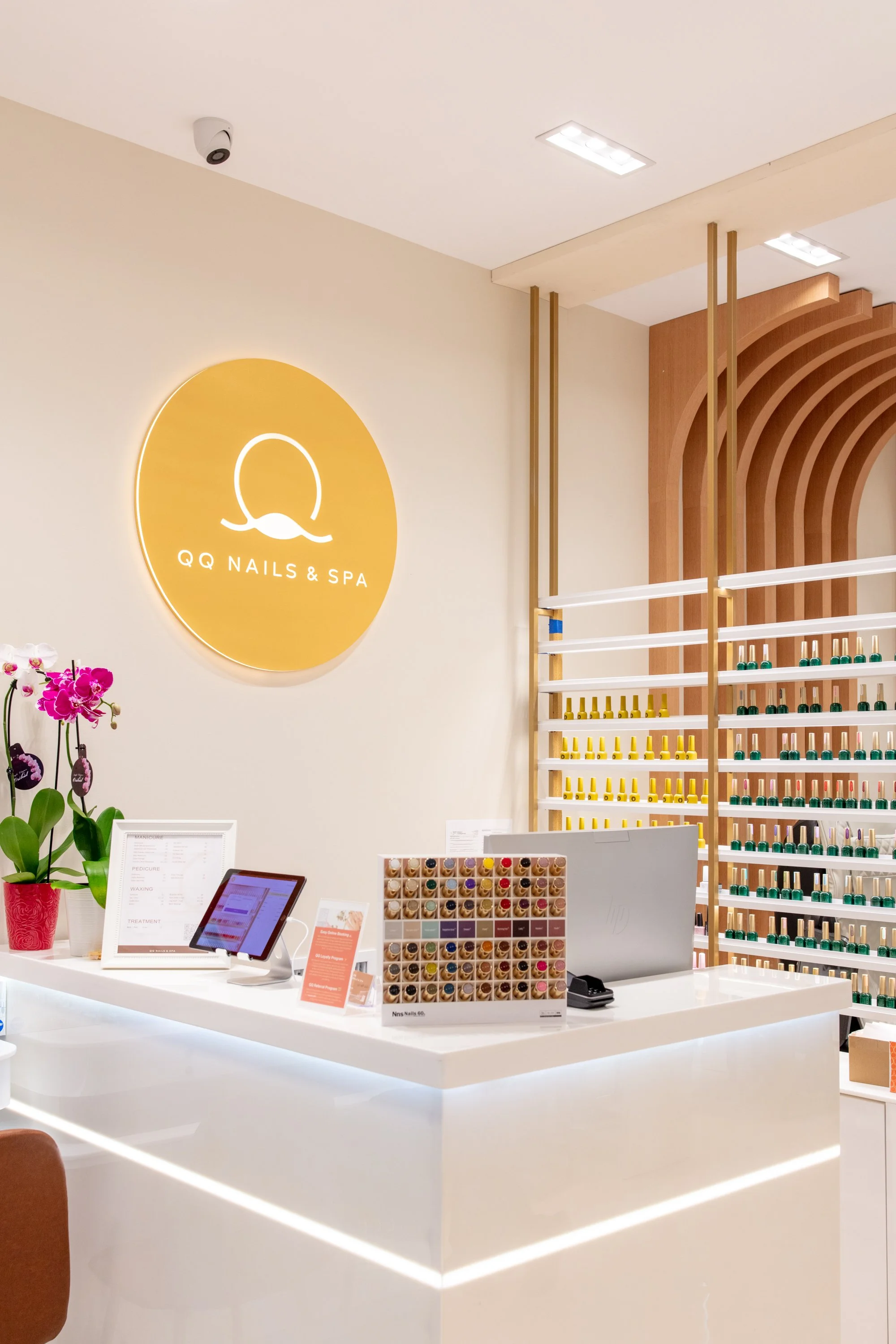 Interior of a nail salon with a white counter, potted orchids, a digital kiosk, sample nail colors, and a back wall with a large yellow logo reading QQ Nails & Spa.