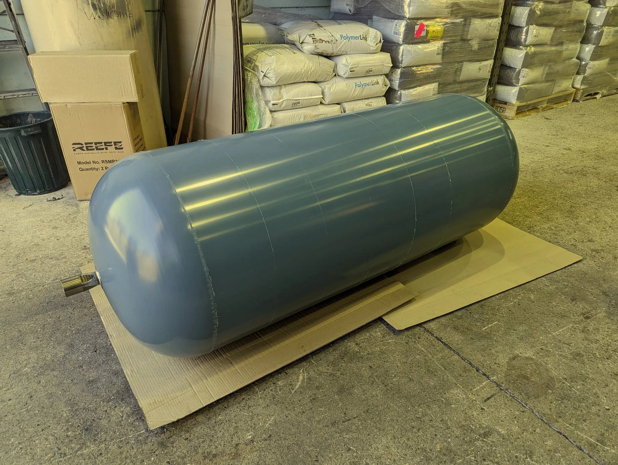 Large gray cylindrical tank on cardboard in a warehouse with packaged materials and a cardboard box nearby.