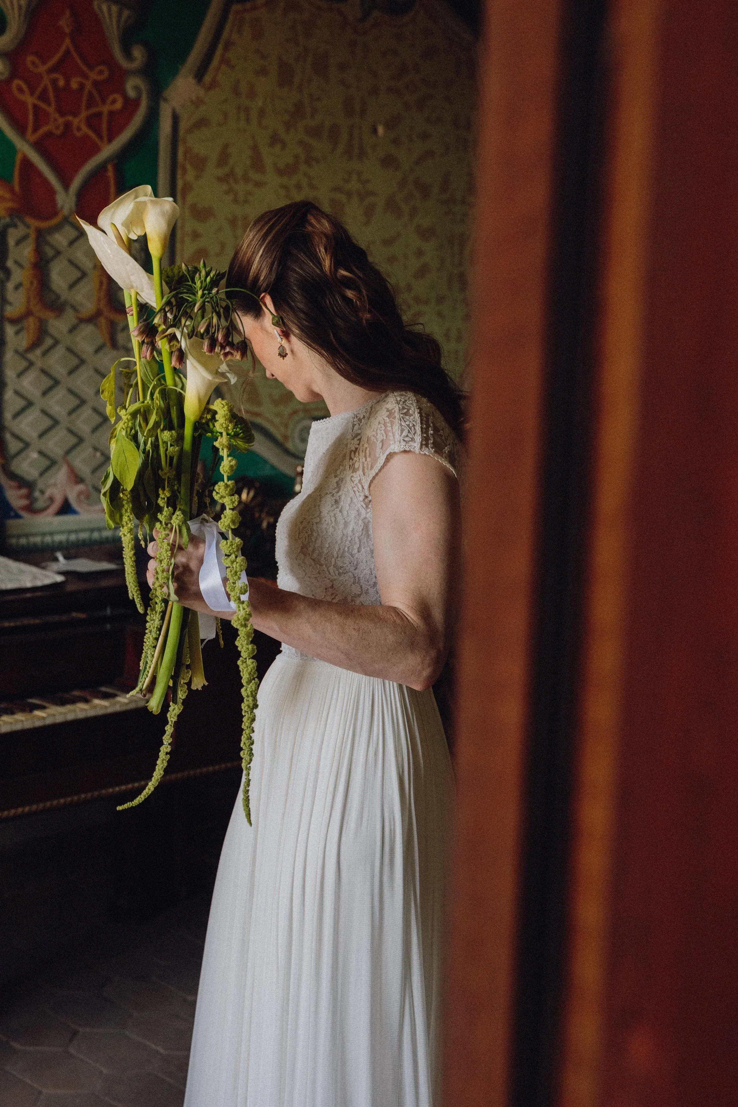 Mujer con vestido blanco, con cabello castaño y rizado, sosteniendo un ramo de flores blancas y verdes, en una habitación decorada con tela y arte antiguo.