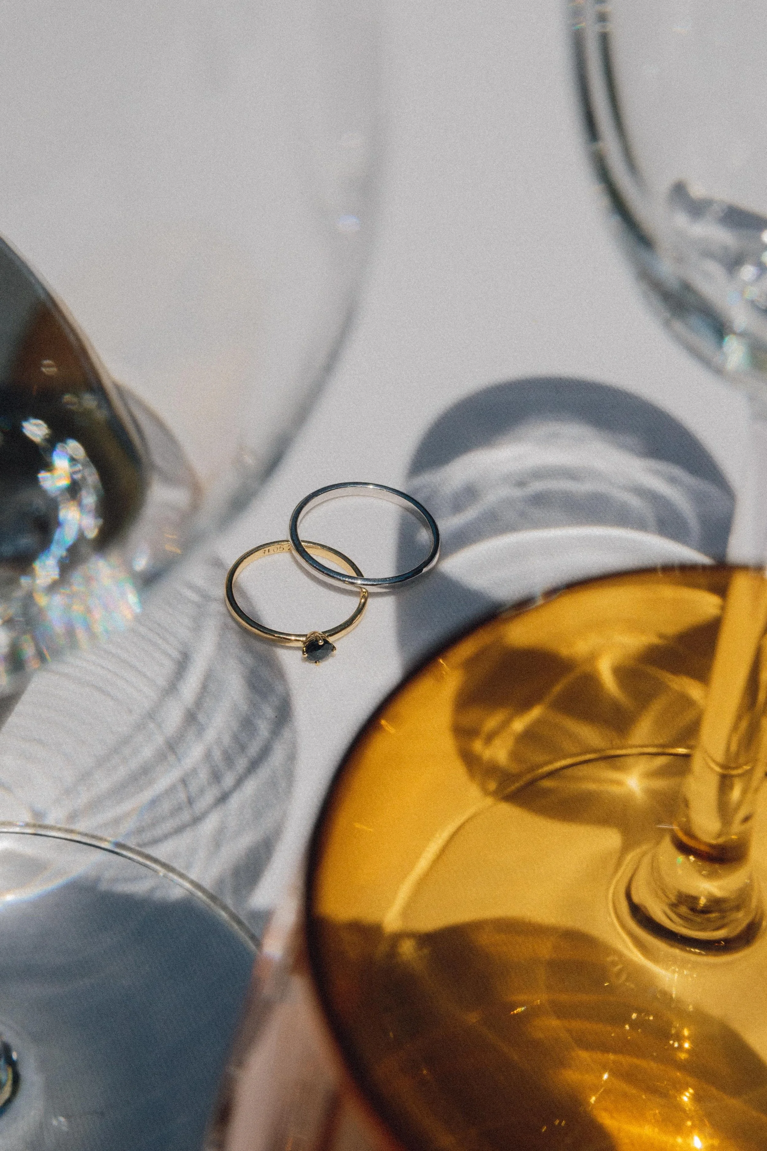 Dos anillos, uno de oro con piedra y uno de plata, en una mesa con copas y vaso con bebida dorada.