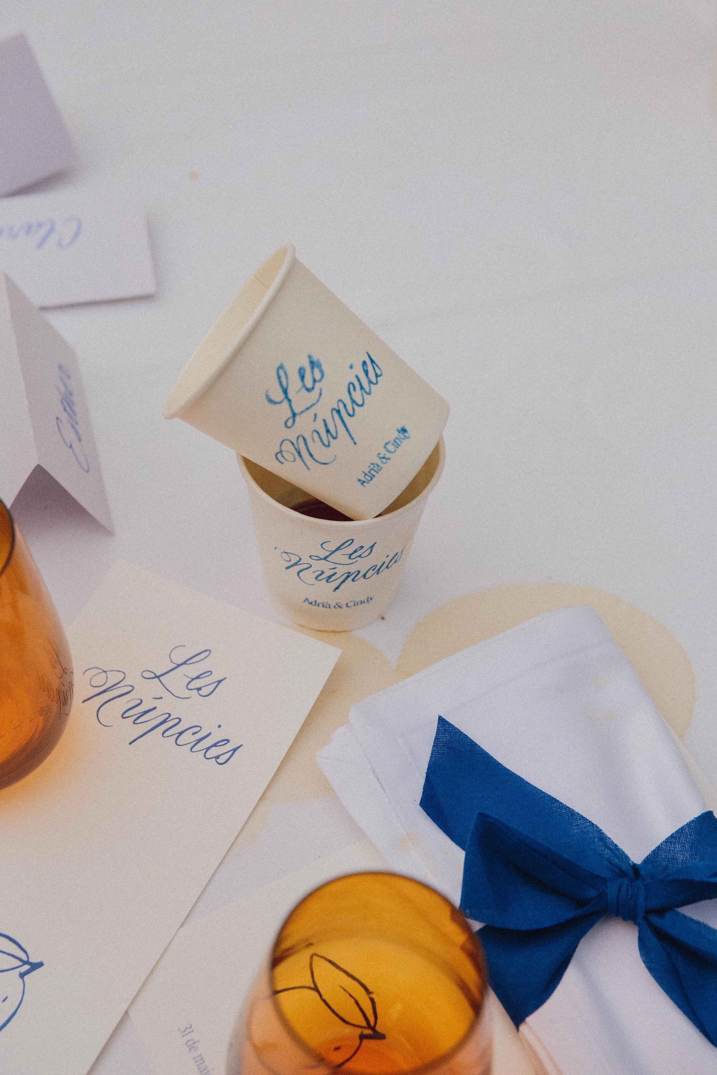 Envases de papel con escritos en letra cursiva y decoraciones en una mesa, con un vaso de cristal anaranjado y un paquete envuelto con lazos azules.
