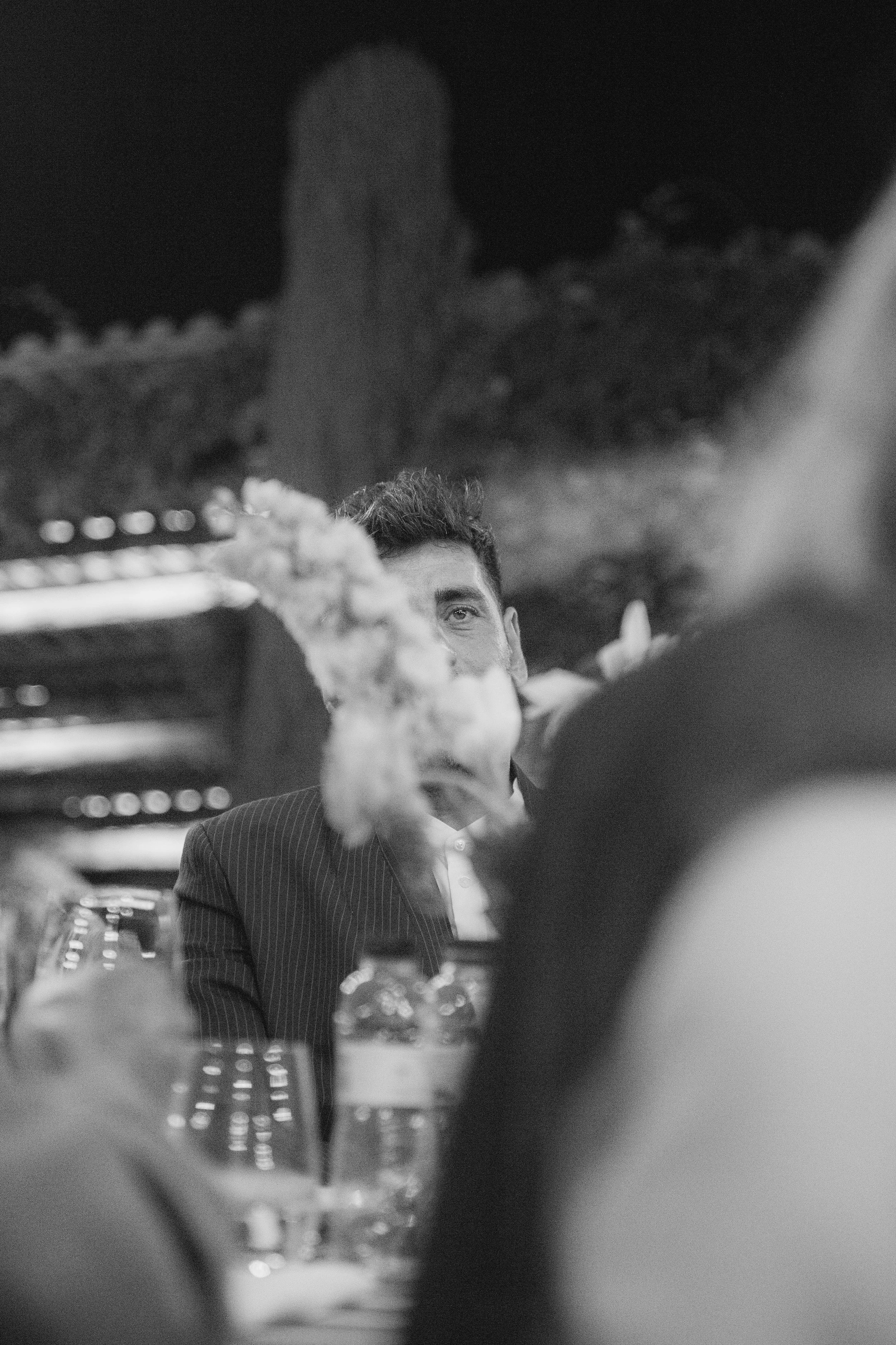 Un hombre con barba y corbata a rayas, sentado en una mesa con botellas de agua, en un entorno que parece un restaurante o evento, con estructuras de fondo y en presencia de varias personas.