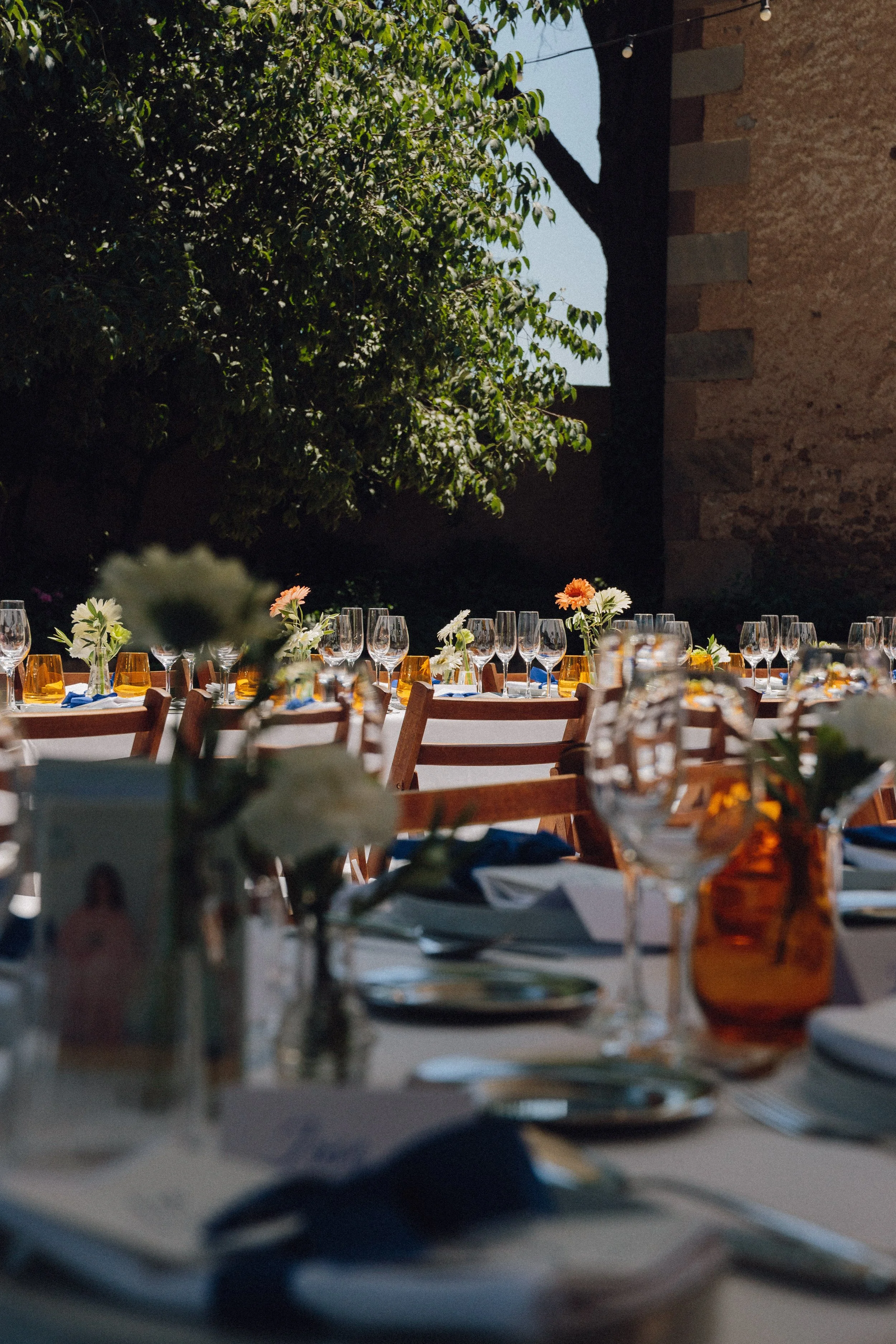 Mesa decorada para una celebración al aire libre con copas, platos, flores y servilletas en un ambiente con sombra de árboles y paredes de piedra.
