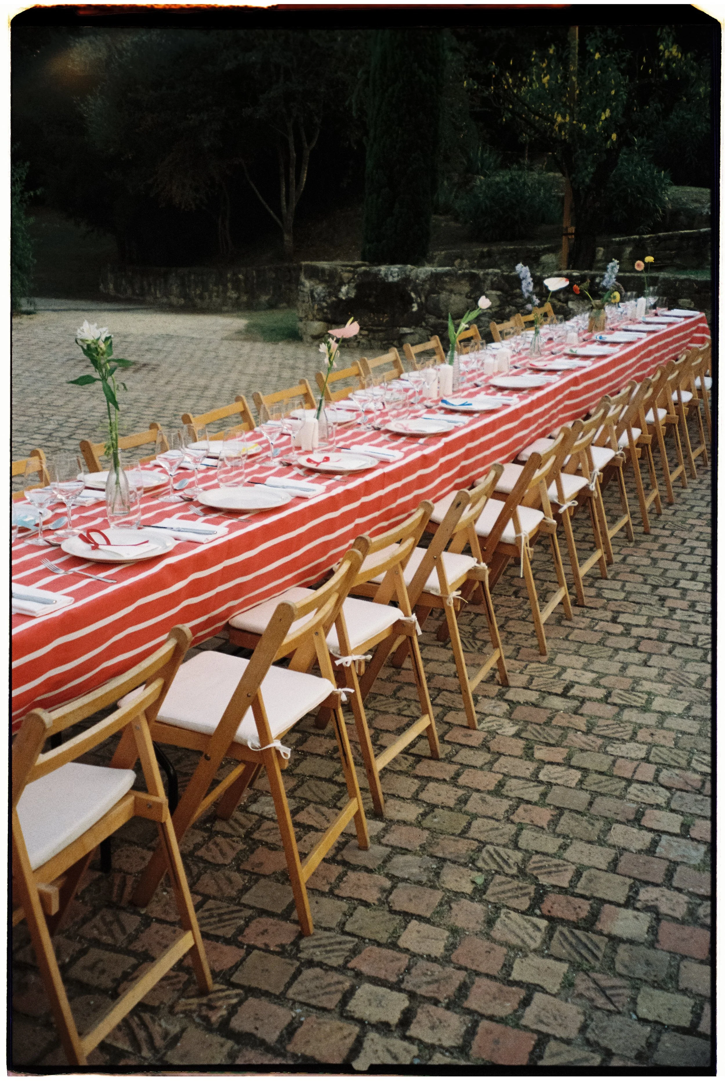 Mesa larga preparada para una comida al aire libre, con mantelería a rayas rojas y blancas, vajilla, vasos y cubiertos, decorada con flores en jarrones, en un entorno exterior por la noche.