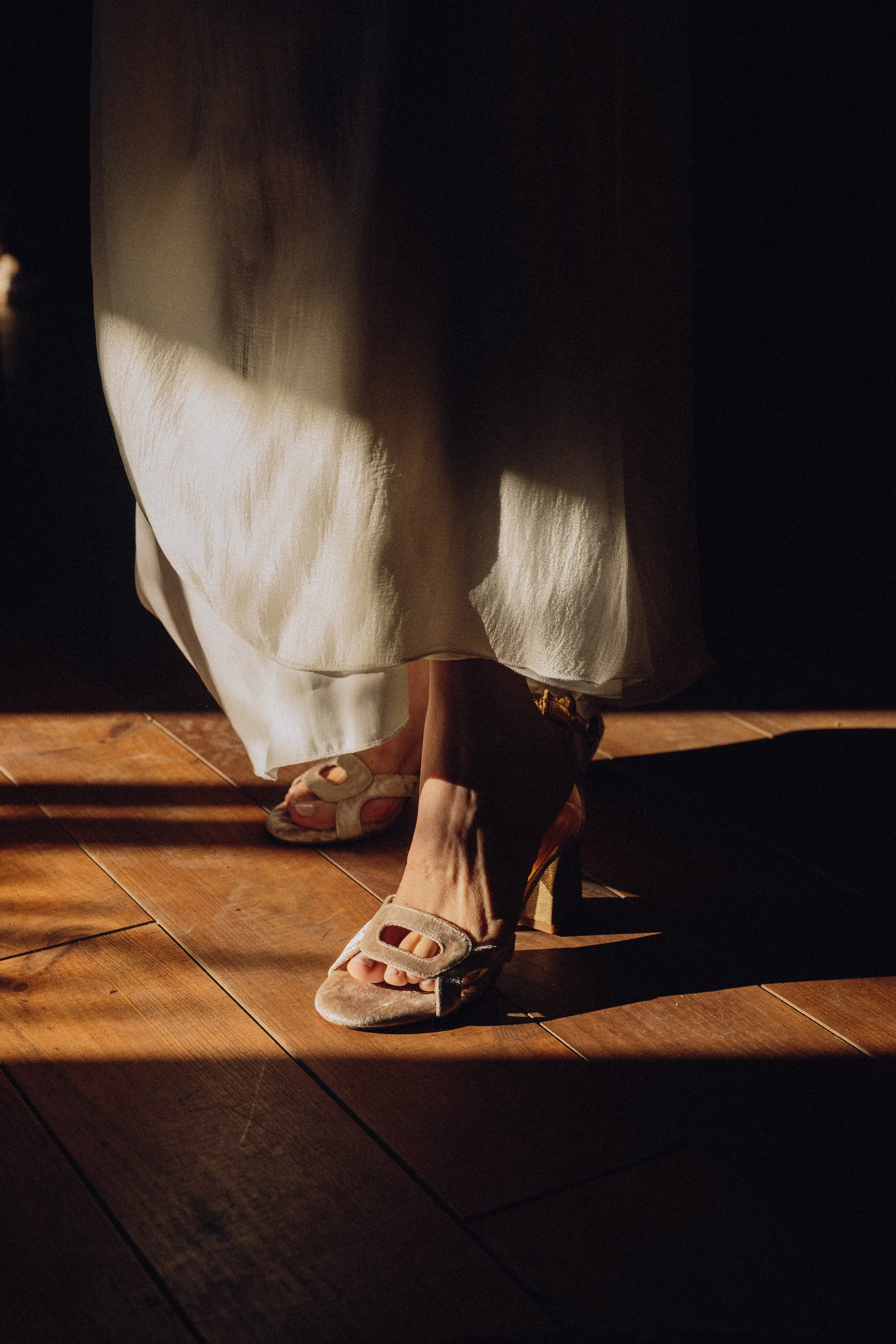 Zapatos de tacón beige con detalles decorativos y vestido largo beige en un suelo de madera, con iluminación y sombra acentuadas.