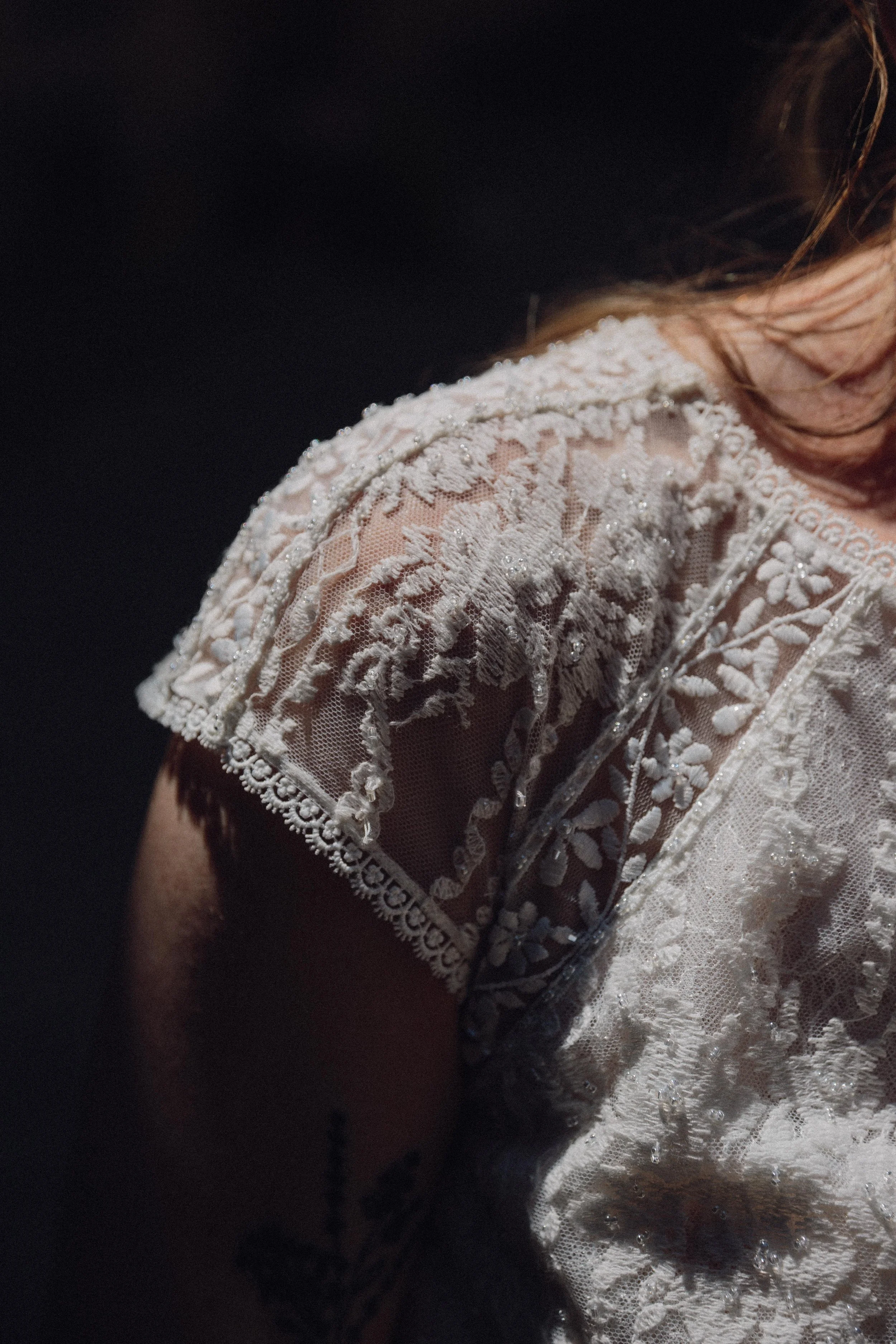 Ropa blanca de encaje con detalles en relieve, mostrando la parte superior del hombro y un tatuaje en el brazo, en un fondo oscuro.