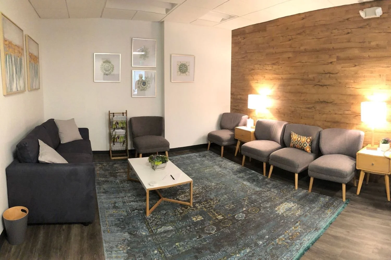 A living room with a black sofa and two matching armchairs, a white coffee table with a plant, a patterned rug, framed artwork on the white wall, and a wooden textured accent wall with two wall-mounted lamps.