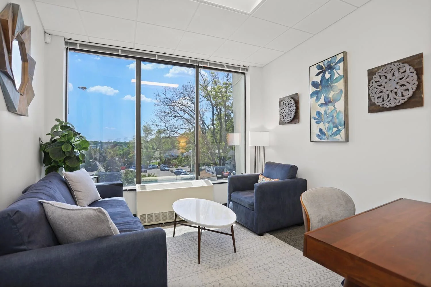 Living room with navy sofas, a white oval coffee table, large window with view of trees and parking lot, wall art with blue leaves and wooden accents, potted plant, and a table lamp.