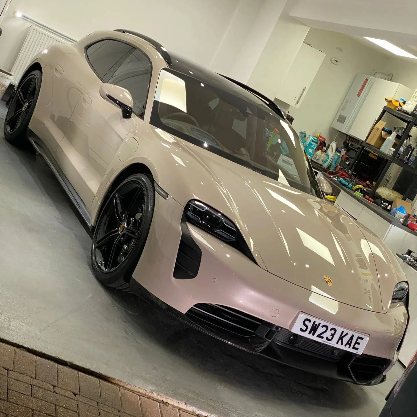 A beige Porsche sports car with black wheels parked inside a garage with shelves and tools visible in the background.