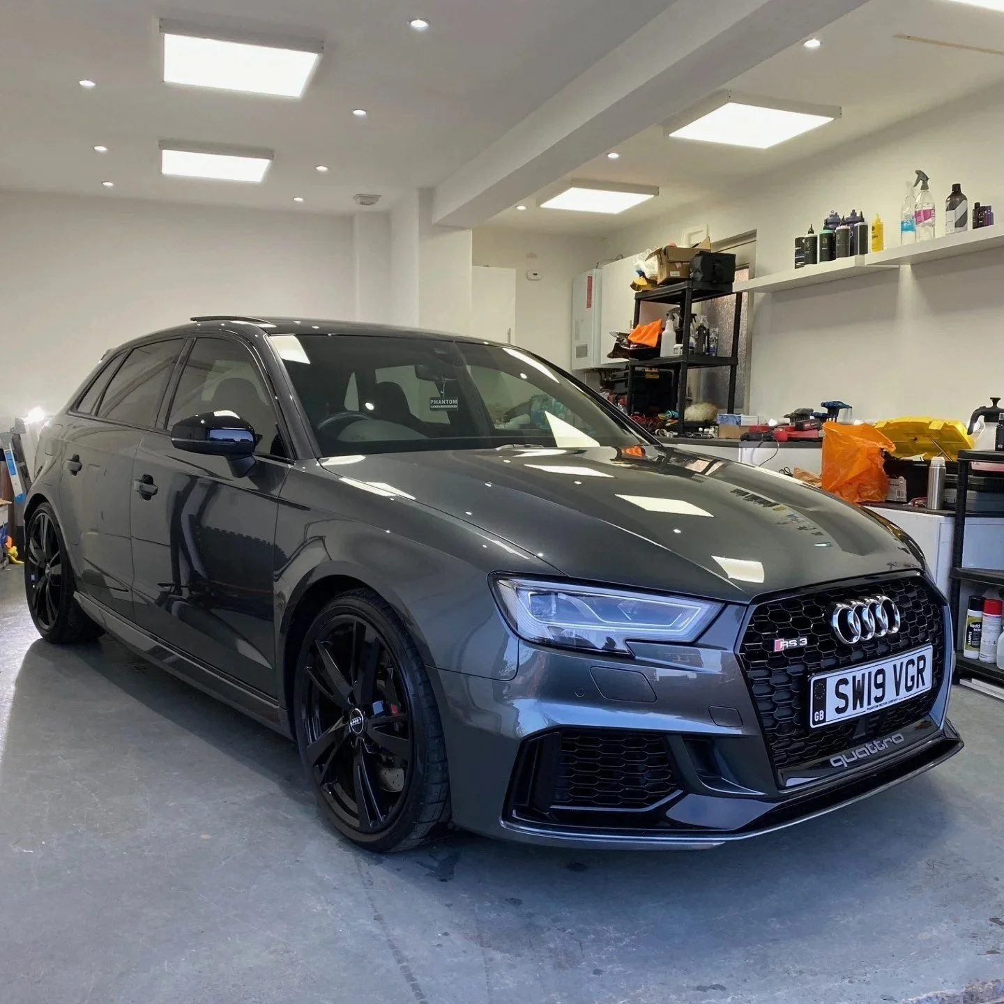 A grey Audi RS3 parked inside a garage with shelves and tools on the right side.