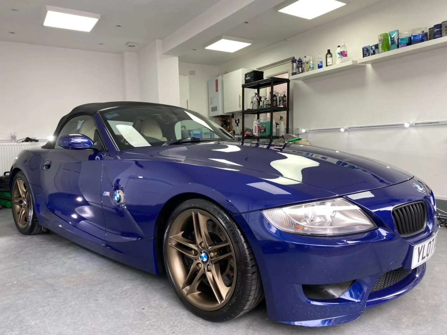 A blue BMW convertible sports car parked in a garage with beige wheels, a soft top, and a BMW emblem. The garage has white walls, a black metal shelf with bottles and other items, and a soft gray floor.