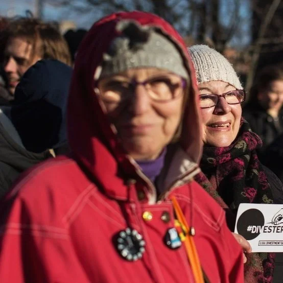 Två vita äldre kvinnor i folksamling utomhus. Den är suddig i förgrunden och tittar in i kameran, den andra ler i bakgrunden.