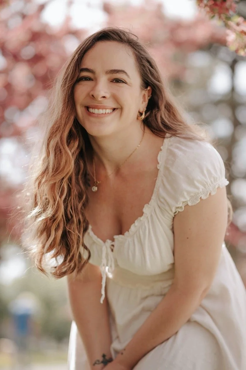 A young woman with long, wavy, auburn hair smiling outdoors during spring or summer, wearing a white dress with puffed sleeves and a v-neckline, with cherry blossom trees in the background.