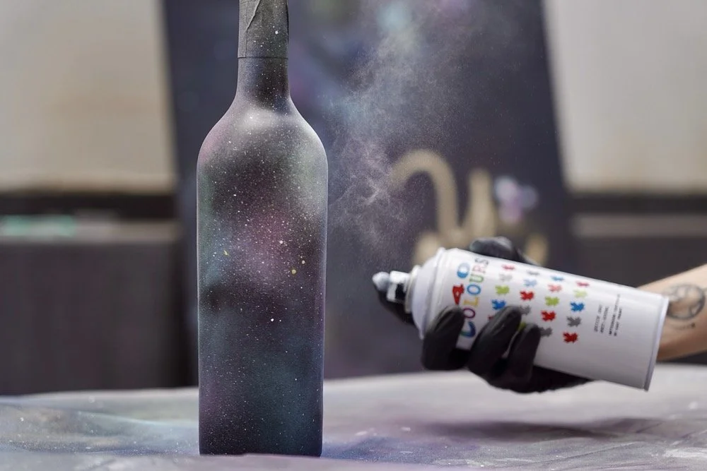 A person wearing black gloves sprays colorful paint onto a black bottle, creating a galaxy-like effect with splatters of paint and spray dust.