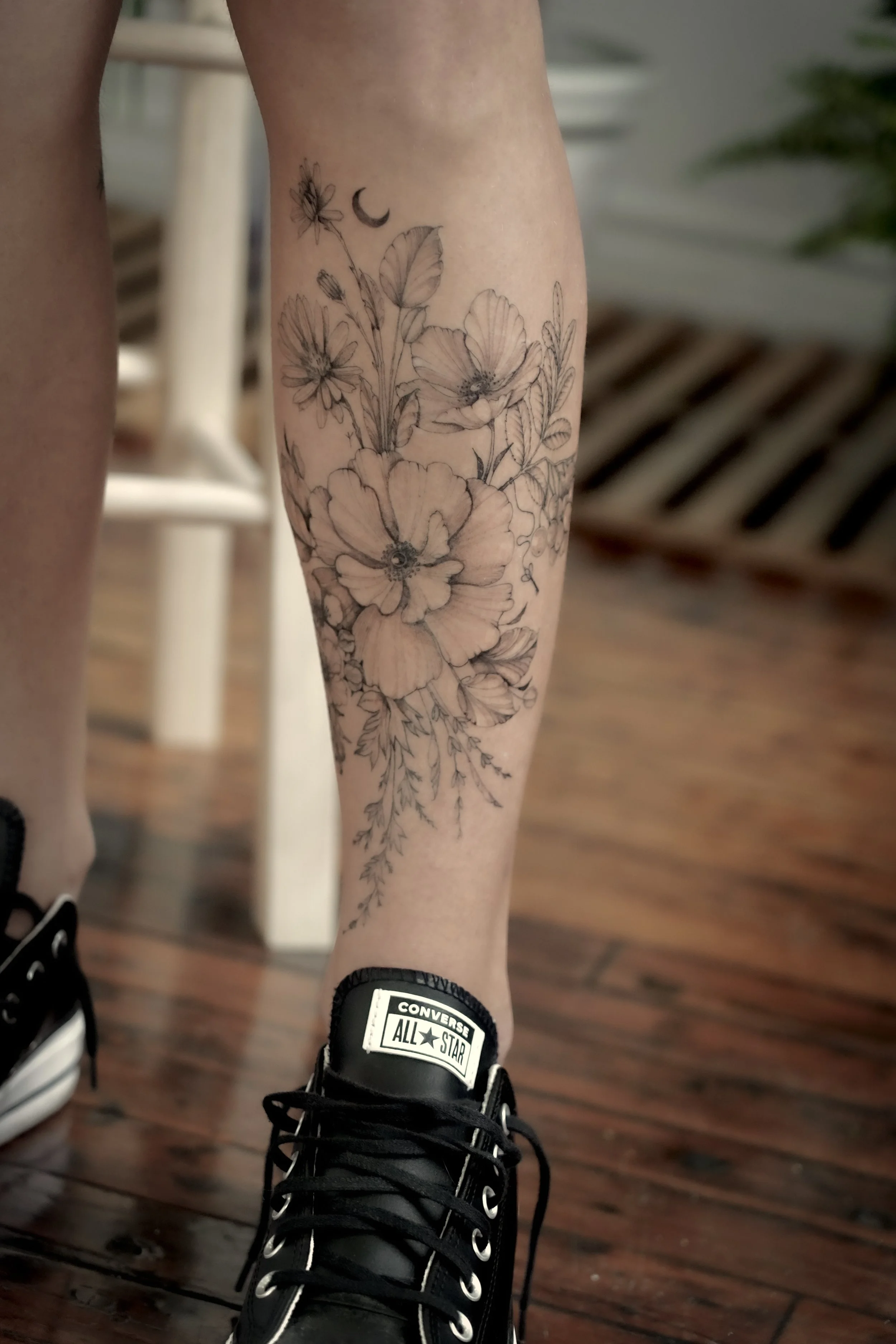 A person's lower leg with a detailed black and gray floral tattoo consisting of large flowers, leaves, a butterfly, and a small crescent moon, done at Inkwaves Tattoo Studio - Montreal