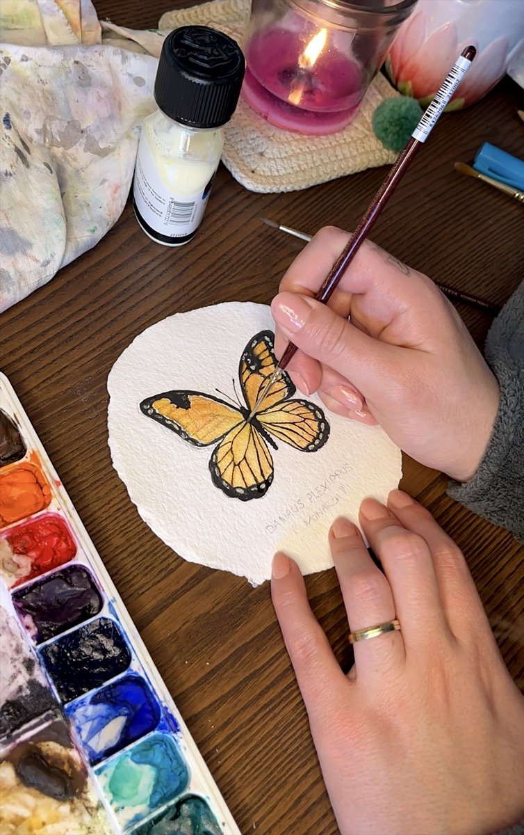 Person painting a watercolor image of a butterfly on textured paper, with watercolor paints and brushes on a wooden table.