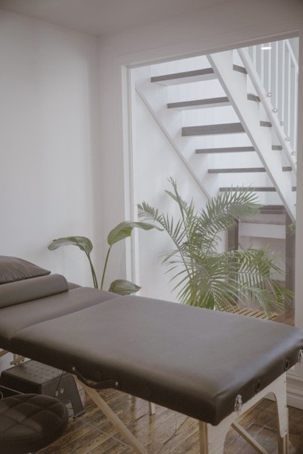 Massage or therapy table placed next to a window with large potted plants outside and a staircase visible through the window.