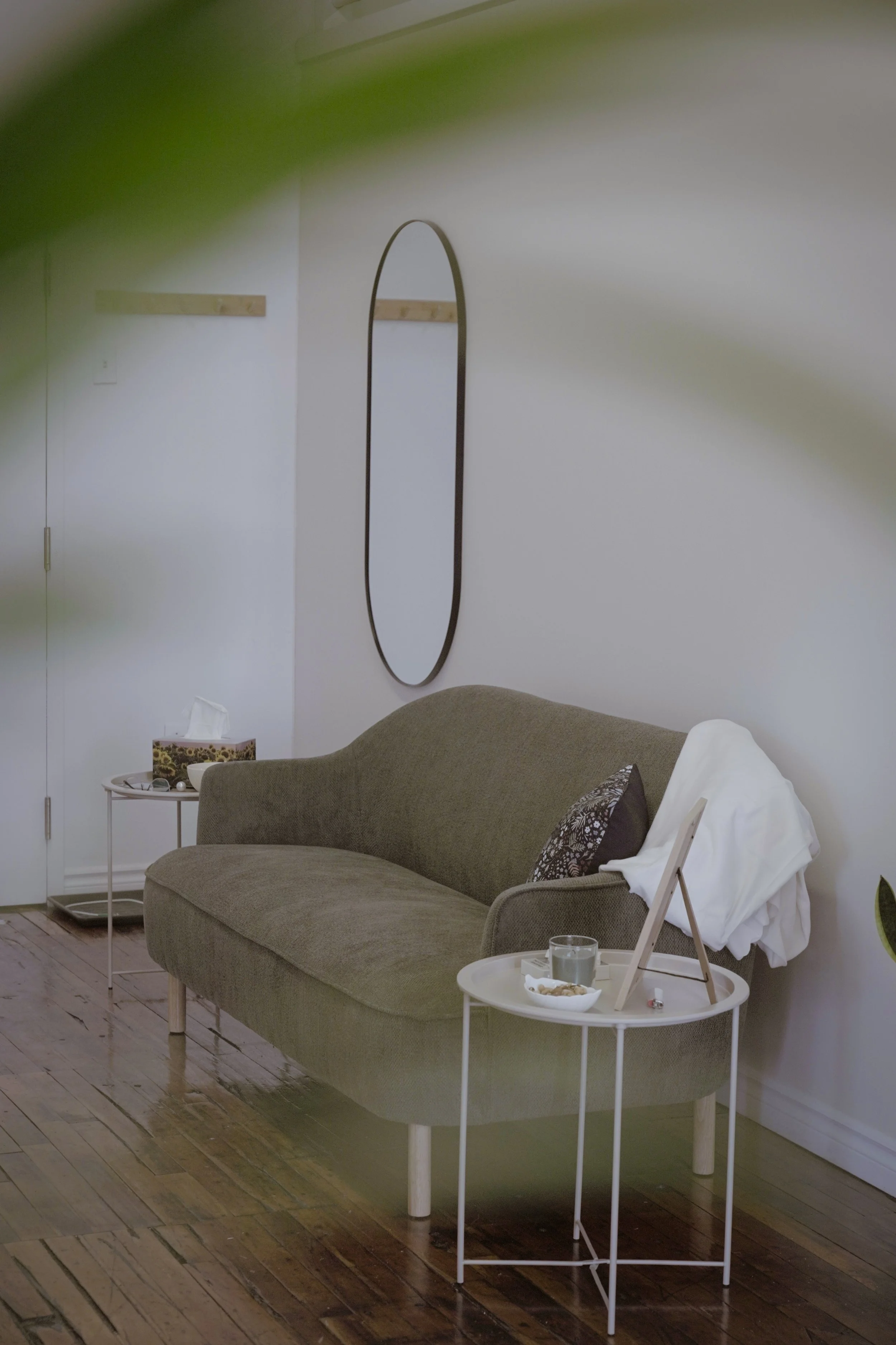 A cozy living room featuring a brown upholstered sofa with patterned pillows, a small white side table with a glass of water and a tray, and a white chair with a white blanket draped over it. Behind the sofa, there's a white wall with a wooden coat r