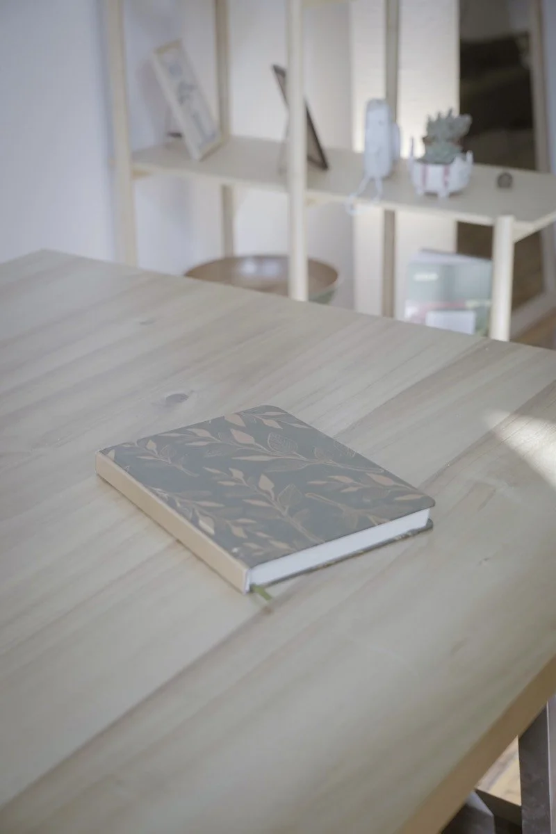 A hardcover book with a leafy pattern on the cover placed on a light wood table.