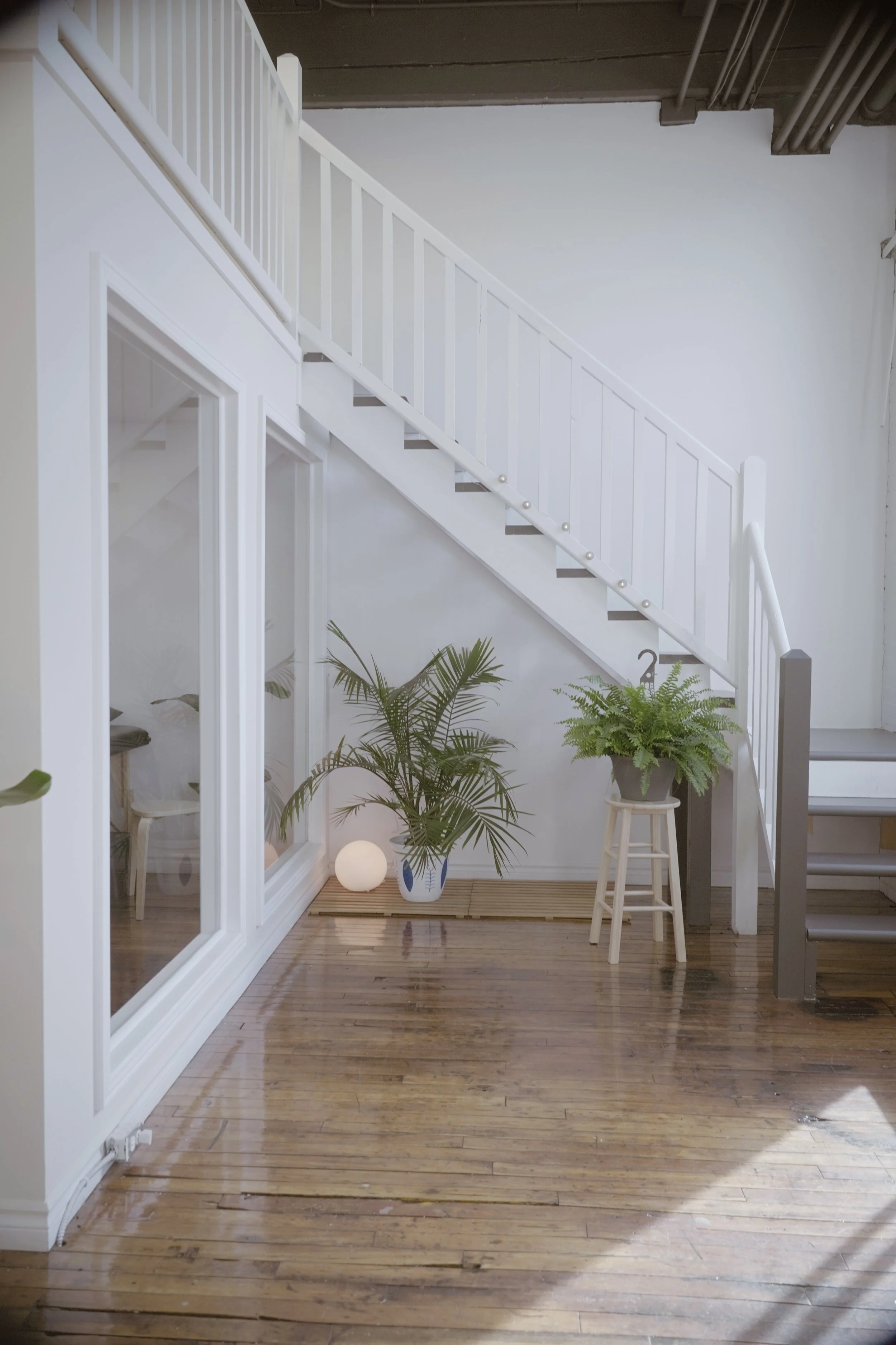 Interior of a modern house with wooden flooring, white walls, a staircase with white railing, and potted plants. There is a small stool, a decorative lamp on the floor, and framed glass doors.