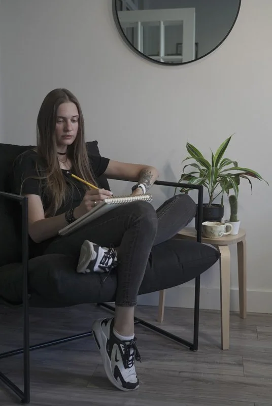 Daria / Inkwaves sitting on a black futon, writing in a notebook with a yellow pencil, wearing black clothes and sneakers, in a room with a round mirror and a potted plant on a small wooden table.