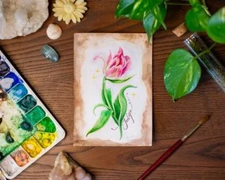 Watercolor painting of a pink tulip on a small canvas, surrounded by green potted plants, watercolor palette, and art supplies on a wooden table.
