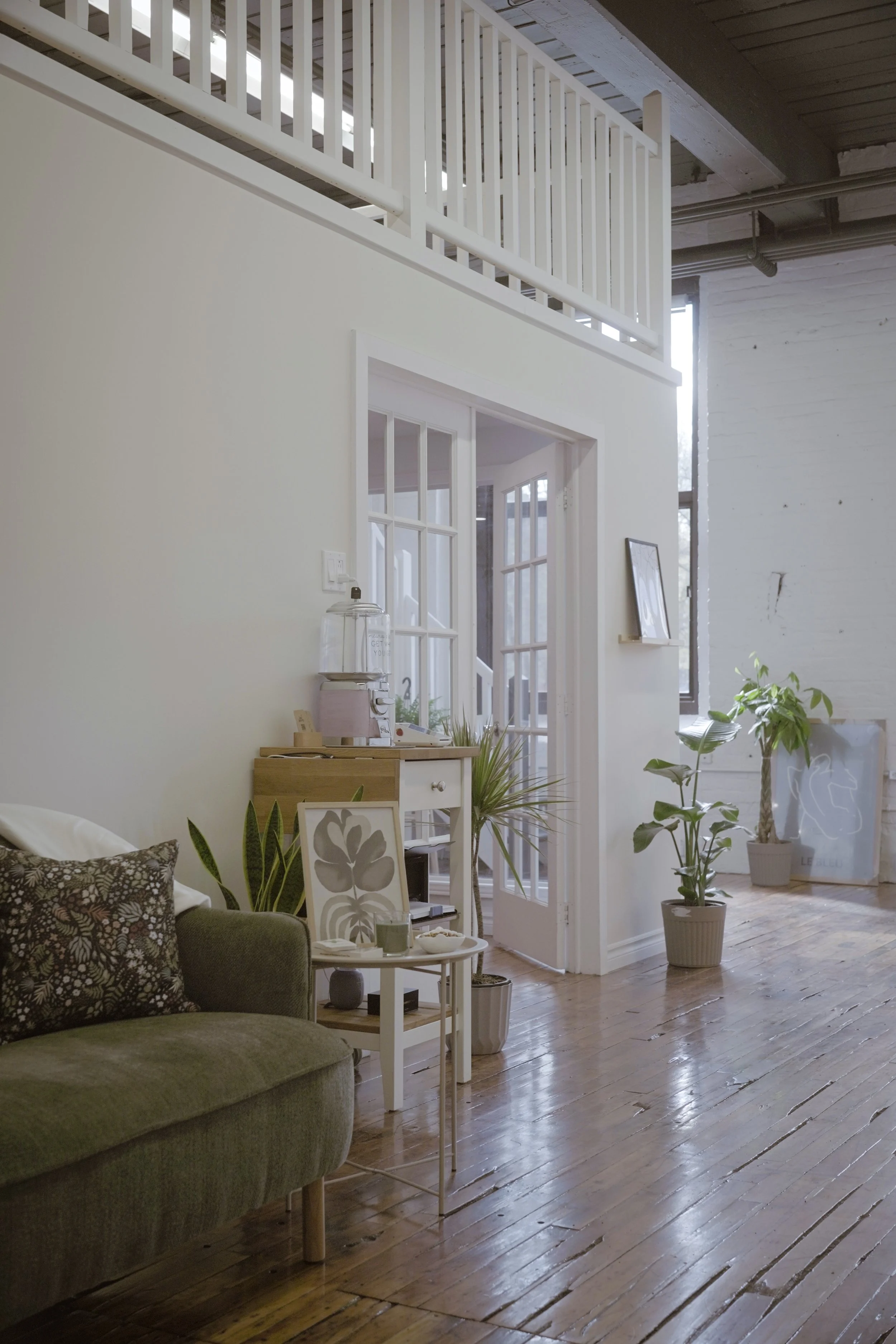 Living room with wooden floor, gray couch with floral pillows, white side table with plants and framed art, and large potted plant near a glass door leading outside. High ceiling with exposed pipes, upper loft with white railing, and large windows le