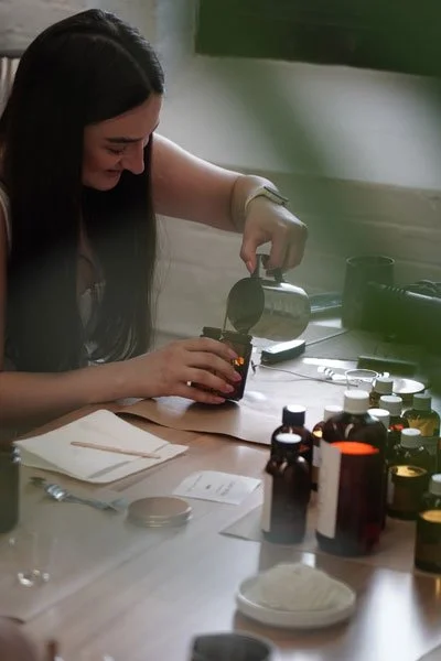 A woman using a spoon to transfer liquid into a small jar on a table with various bottles and containers.