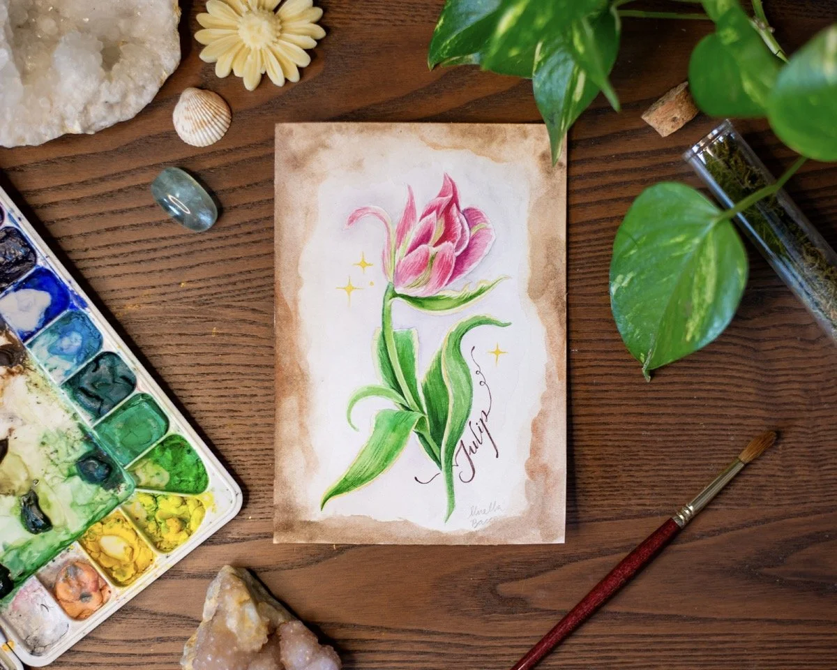 A watercolor painting of a pink tulip flower with green leaves, surrounded by yellow sparkles, on a brown and white background, placed on a wooden surface with various art supplies and decorative items around.